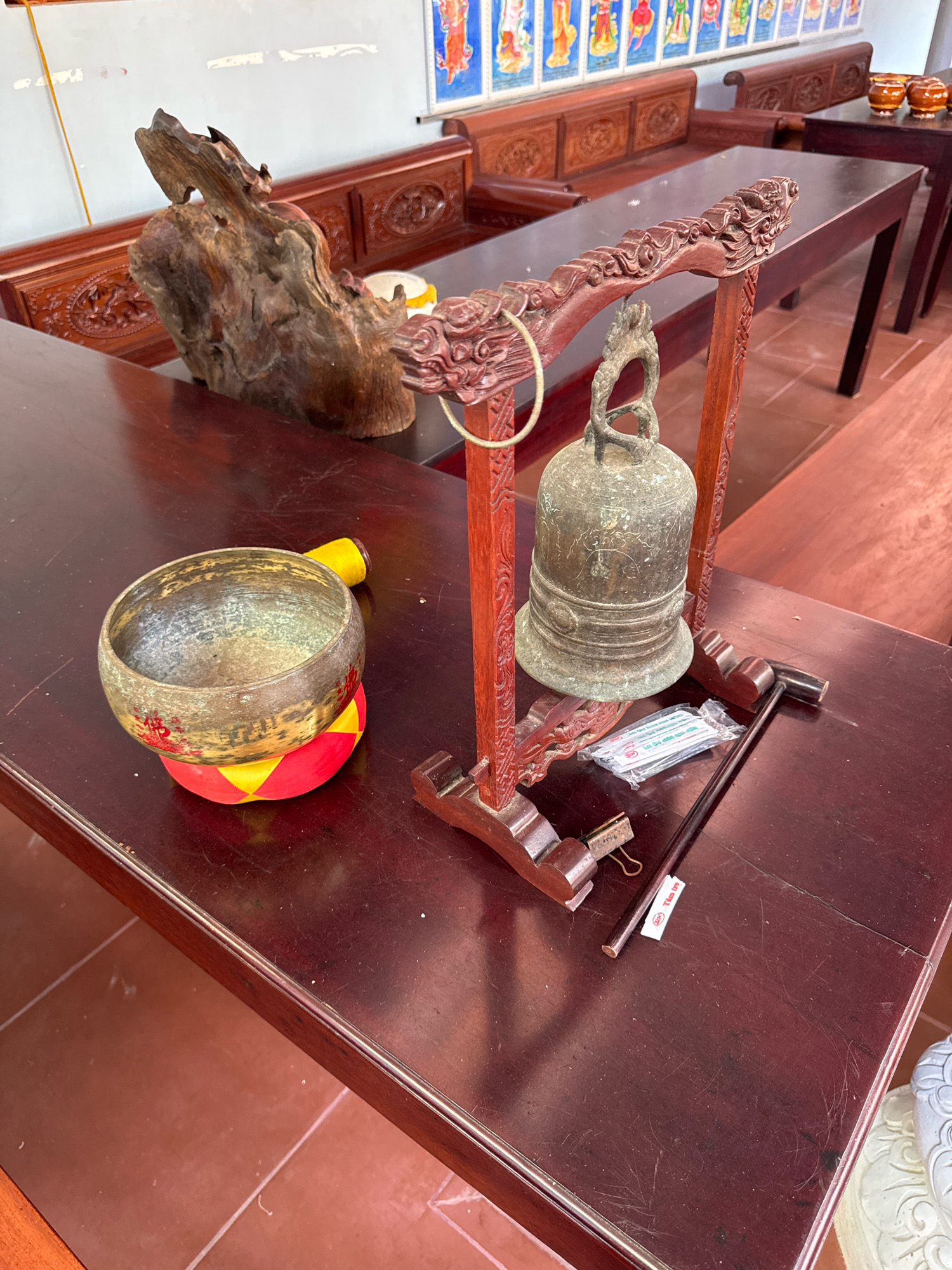 The image depicts a close-up, high-angle view of a wooden table in what appears to be a Buddhist temple or religious establishment in Nha Trang, Vietnam. On the table, there is a collection of objects, including a large, antique bronze bell hanging from a carved wooden stand that is adorned with dragon motifs. To the left of the bell is a brass singing bowl, resting on a colorful cushion. Adjacent to the bell and stand, a wooden mallet with a black handle lies horizontally on the table, alongside a small packet of what appear to be incense sticks or similar items. A piece of weathered, organic wood sculpture is positioned to the left of the singing bowl. The table itself is made of dark, polished wood and is set against a backdrop of more dark wooden furniture, possibly benches, with ornate carvings. The wall behind the furniture is light-colored, and a colorful, framed artwork featuring multiple figures is visible high on the wall. The floor is tiled in a reddish-brown hue. The lighting suggests an indoor setting, possibly with natural light filtering in. There are no people visible in the frame. The overall scene conveys a sense of cultural and spiritual significance.