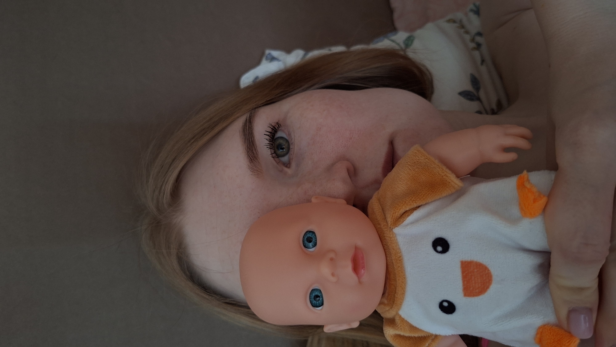 A woman with fair skin and freckles is lying down next to a pillow with a floral pattern, holding a doll. The doll has a bald head, blue eyes, and is wearing a white outfit with orange accents. The scene appears to be indoors. The location is Chaikovsky, Russia.