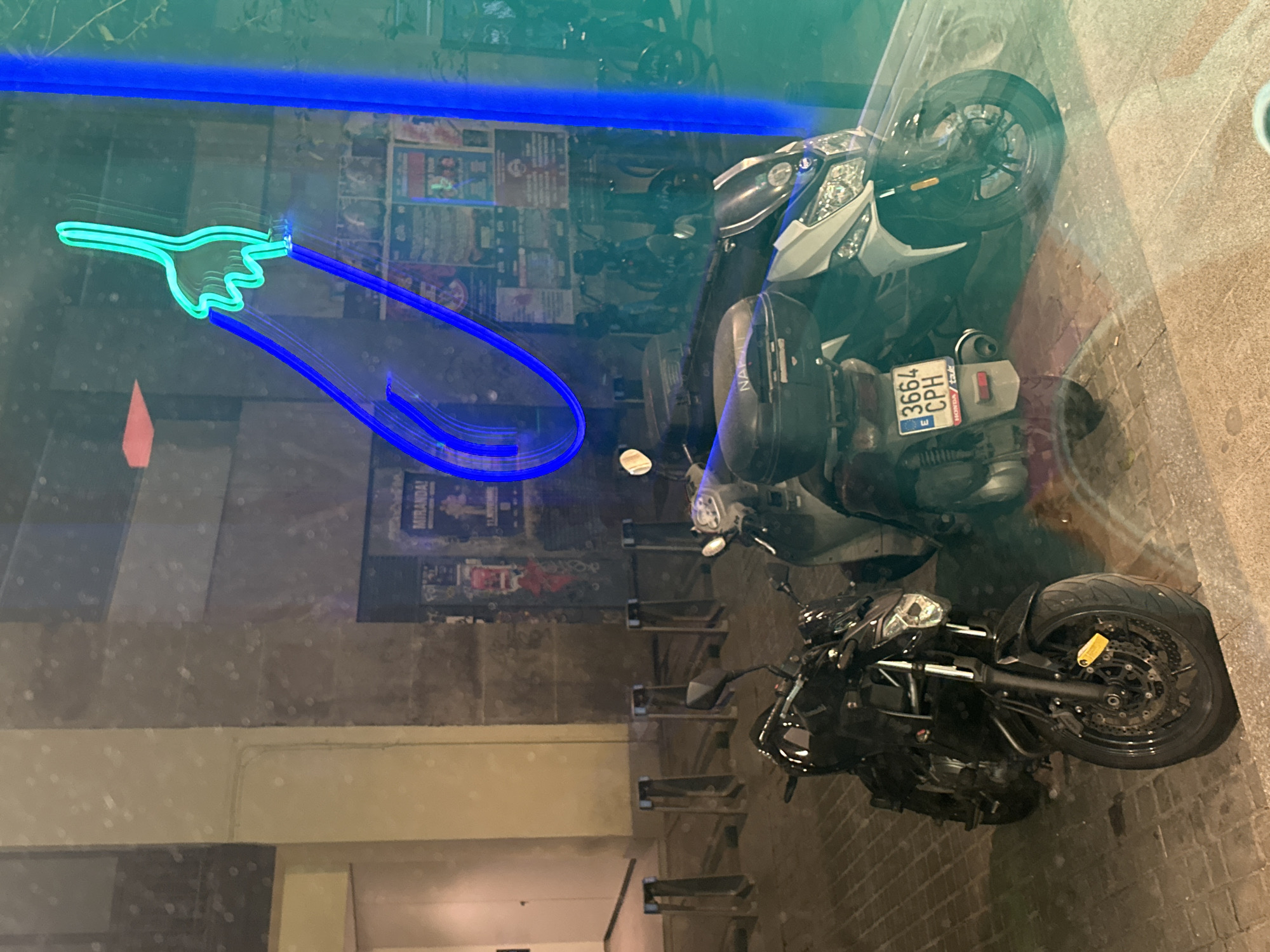 The image depicts a street scene in Madrid, Spain, viewed through a glass window with a neon sign of a pointing hand. Three motorcycles are parked along the sidewalk. The motorcycles appear to vary in style and size. The background shows posters, adding to the urban setting. The scene is generally dark, illuminated by streetlights and the neon sign.
