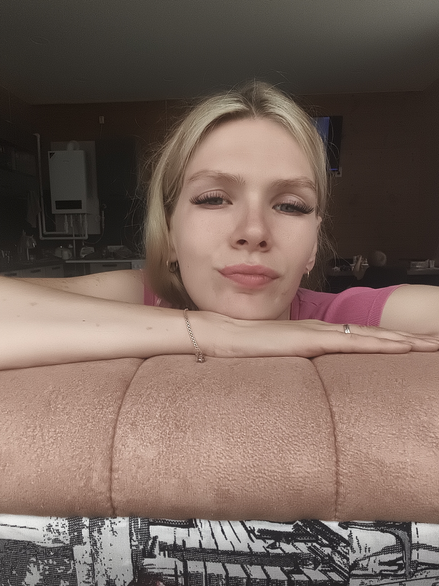 A young woman with blonde hair and long false eyelashes rests her head on her arms, which are placed on a textured pink surface. She is wearing a pink top and a bracelet with a charm. A ring is visible on her right hand. The background is dark and indistinct, with a white appliance mounted on the wall and what appears to be a television screen visible on the right. The scene is indoors, possibly in a home. The location is identified as Русский Вожой, Russia.