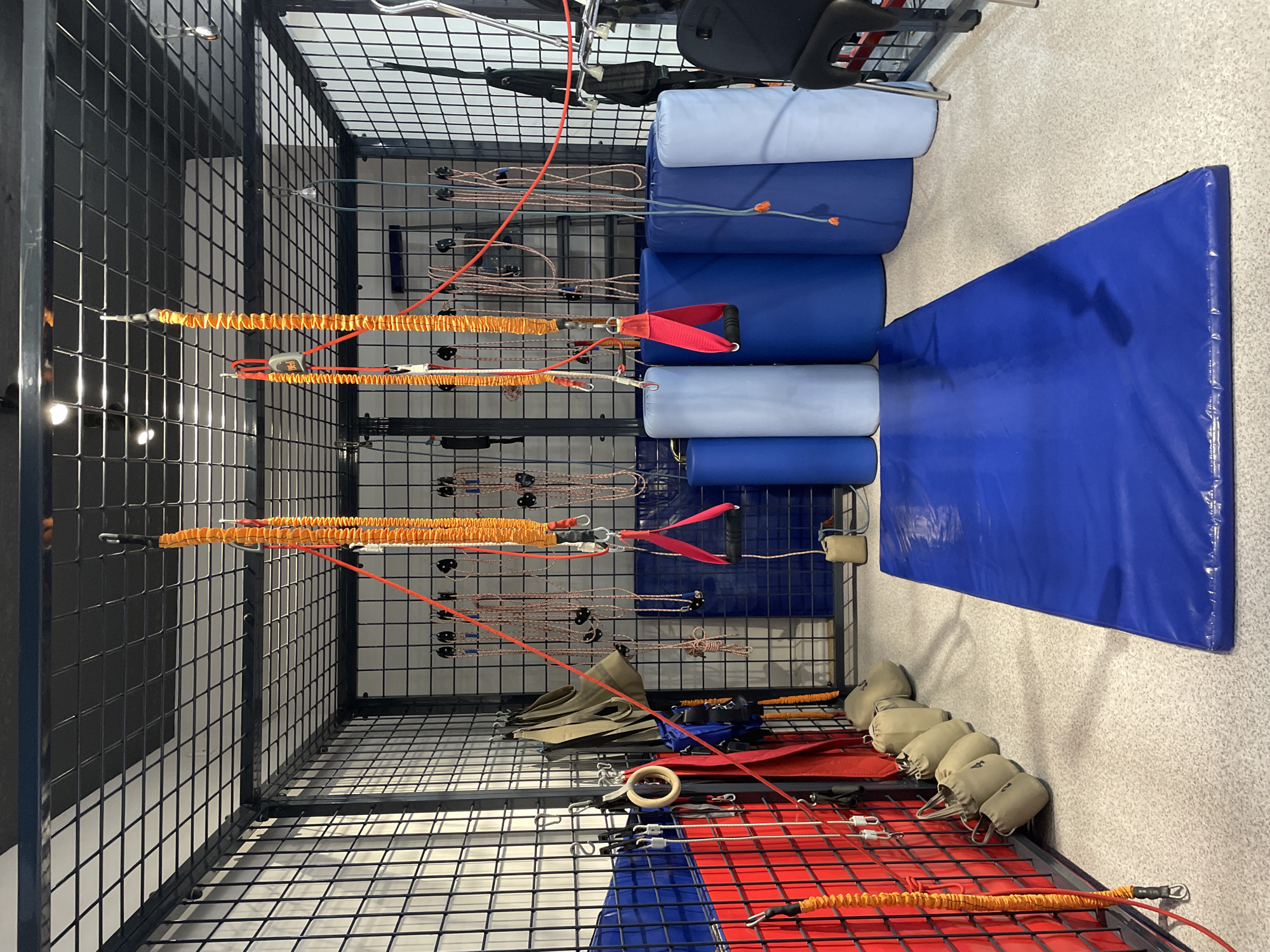 The image shows the interior of a gym, specifically a cage-like structure with various exercise equipment. Hanging from the ceiling are orange resistance bands with red handles, red ropes with black grips, and other suspension training apparatus. The walls are made of wire mesh, and the floor is a speckled grey material. Against the back wall are several large, cylindrical blue and light blue mats, and in the foreground, a large blue mat lies on the floor. There are also several sandbags hanging from hooks on the wall. The lighting suggests an indoor environment, possibly a fitness studio or a specialized training facility. The image does not contain any identifiable landmarks or specific architectural features that would allow for pinpointing a location within Almaty, Kazakhstan.
