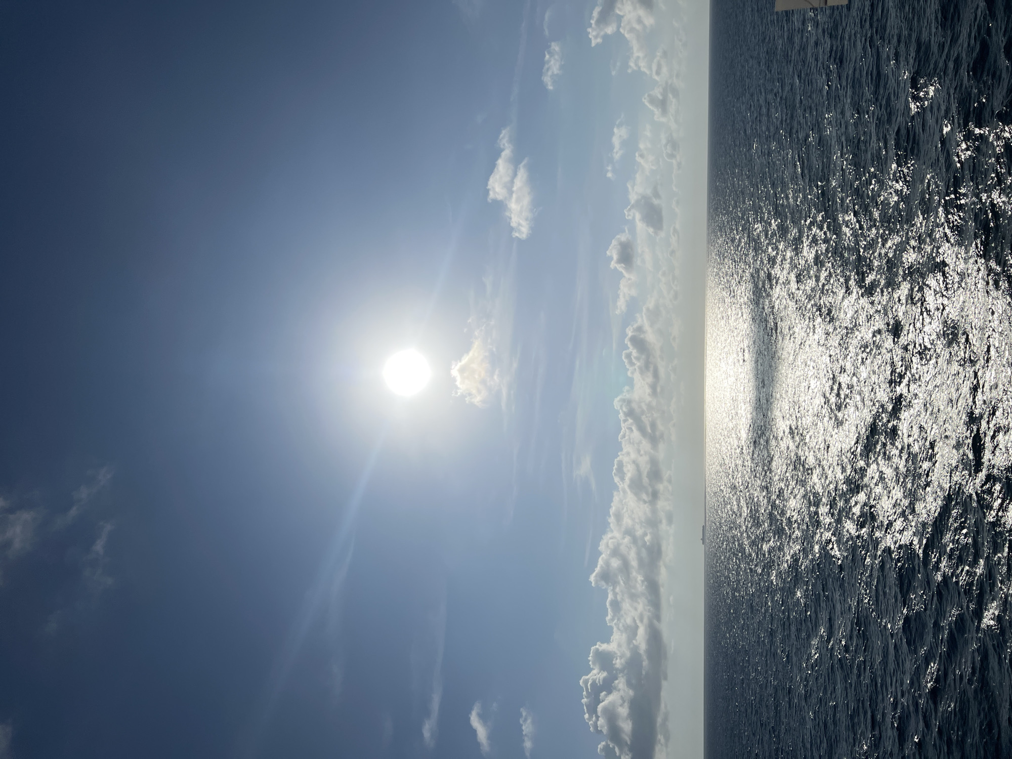 A wide shot captures a sunlit expanse of the ocean under a partly cloudy sky. The sun, a bright orb, is positioned high in the sky, casting a shimmering reflection across the dark blue water. Scattered clouds, some fluffy and white, others grayer, dot the horizon. In the far distance, a ship is visible as a small silhouette against the bright sky. The water is textured with ripples and small waves, indicating a gentle movement of the sea. The overall scene conveys a sense of open space and the vastness of the ocean.