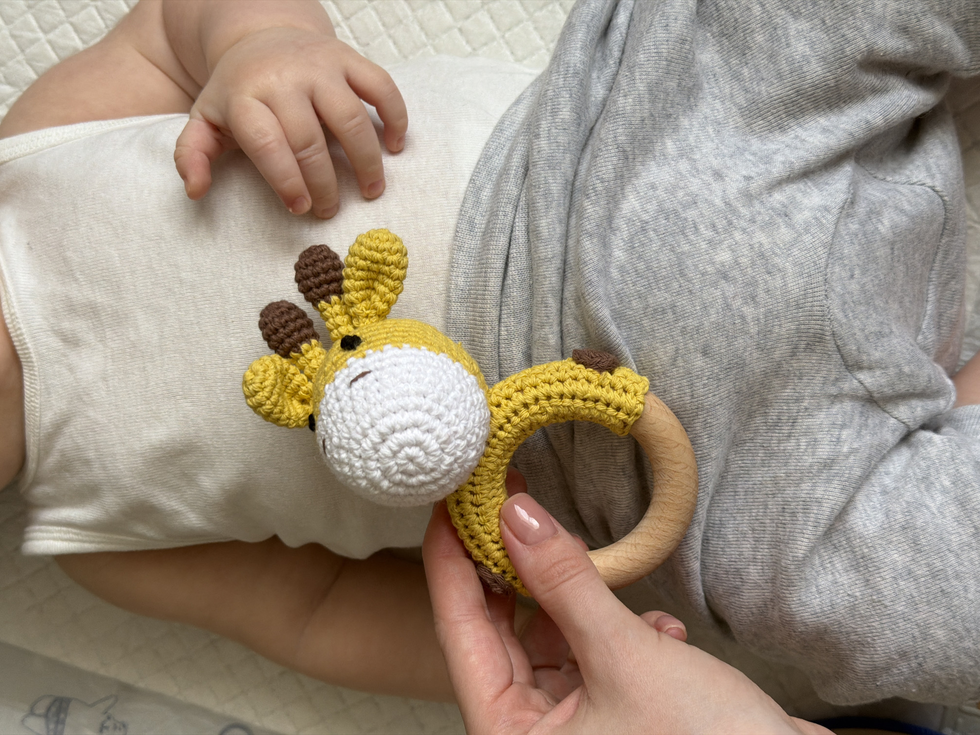 An overhead shot shows a baby lying down, partially dressed in a white bodysuit and light grey bottoms, with an adult hand presenting a crocheted giraffe rattle. The baby's tiny hand is visible near the top left, resting on the white bodysuit. The crocheted toy features a yellow body and head with a white snout and brown ears/horns, attached to a smooth, light wooden ring. The adult's hand, with neatly manicured nails, holds the wooden ring of the toy, offering it towards the baby. The background suggests a soft, quilted white surface, likely a blanket or mat. The scene depicts an interaction between an adult and a baby, possibly playing or comforting, in an indoor setting in Khimki, Russia. No visible text is present. The time of day and weather cannot be determined.
