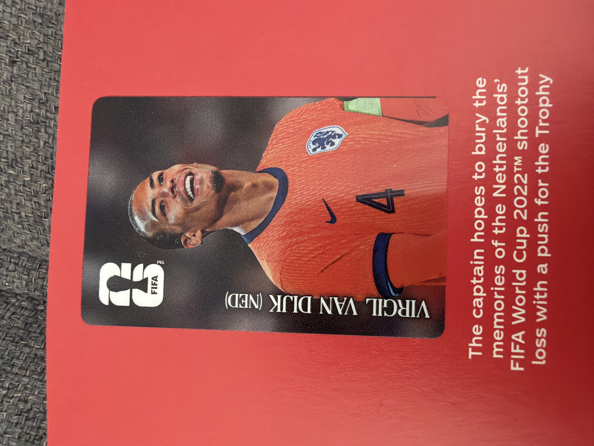 The image shows a close-up of a FIFA trading card featuring a portrait of a smiling man in an orange jersey with the number 4 on it. The man is likely a soccer player. The card has the FIFA logo and the name "VIRGIL VAN DIJK (NED)" printed vertically on the left side. Below the card, white text reads "The captain hopes to bury the memories of the Netherlands' FIFA World Cup 2022™ shootout loss with a push for the Trophy". The background is a solid red surface. There is no visible indication of the city of Zlatitrap, Bulgaria, or any elements that would place the image in that specific location.