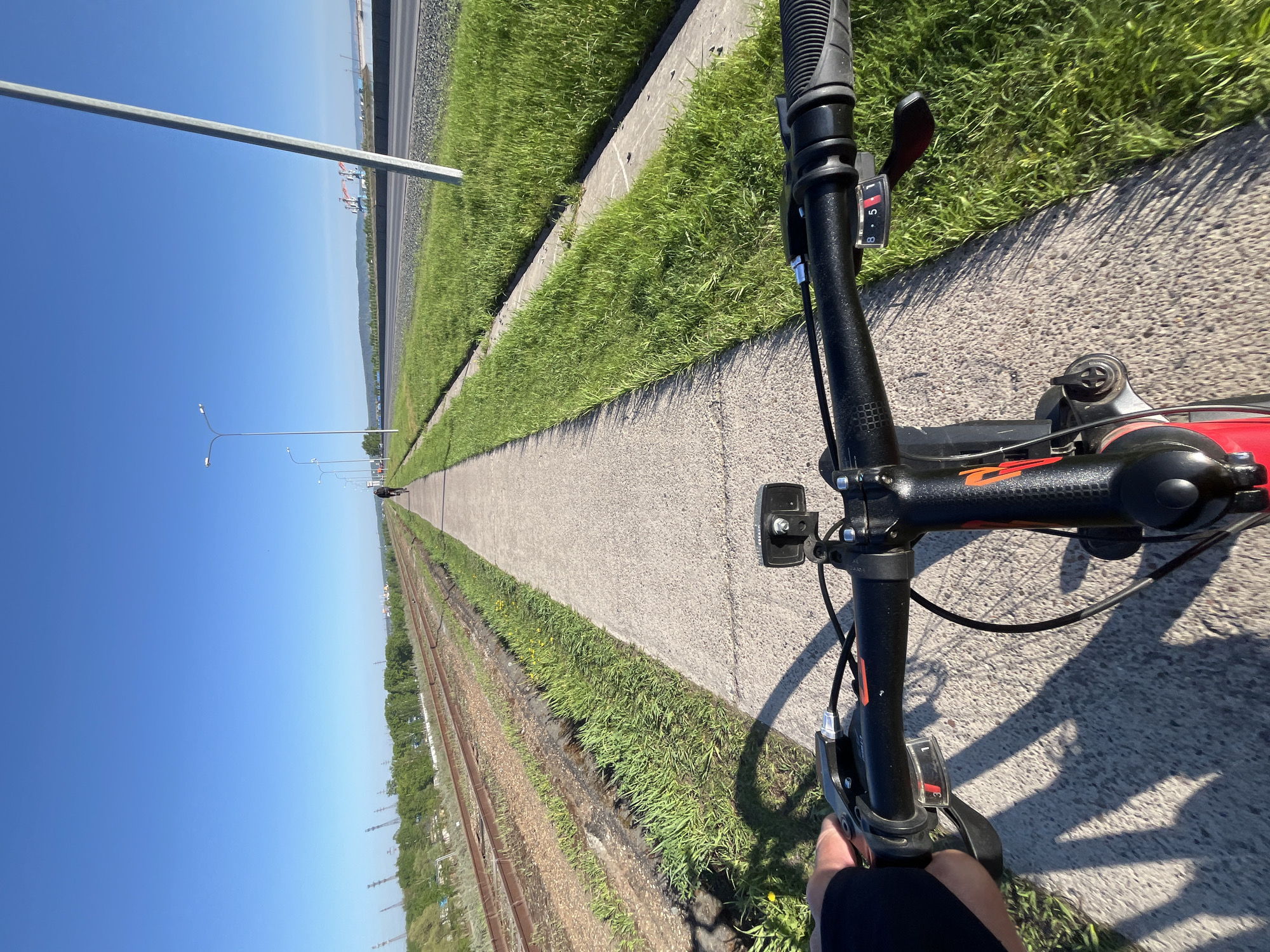 The image shows a first-person perspective of a bicycle ride on a paved path adjacent to a set of railway tracks. The path is bordered by grass on both sides. In the distance, the path appears to narrow due to perspective, leading towards a horizon with industrial structures. Lampposts line the path further away. The sky is clear and blue. The bicycle's handlebars, gear shifter, and part of the frame are visible in the foreground.
Based on the industrial structures and the railway, this is likely somewhere in or near the industrial zones of Chaikovsky, Russia. The image shows clear, sunny conditions.
