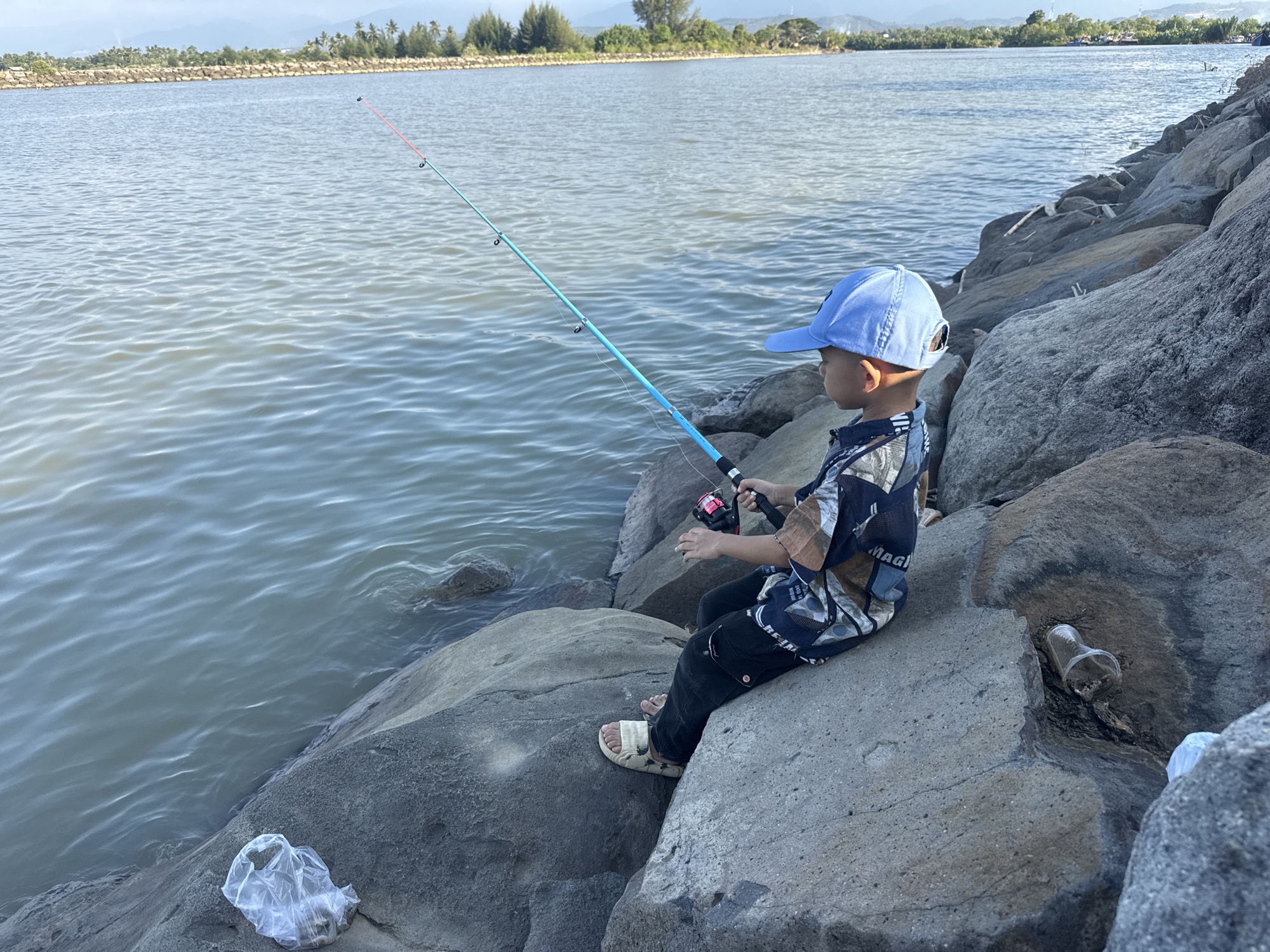 A young boy wearing a blue baseball cap and a patterned shirt sits on large, gray rocks along the edge of a body of water. He is holding a fishing rod, which has a light blue colored shaft with red accents near the tip, and a red fishing reel. The boy is looking out at the water. The water is calm with small ripples. In the background, a shoreline with trees and some buildings can be seen under a hazy sky. There are other rocks scattered around the boy, and some discarded plastic items, including a plastic bag and a clear plastic cup, are visible on the rocks. The scene appears to be located at the coast of Reudeuep, Indonesia, likely a fishing spot.