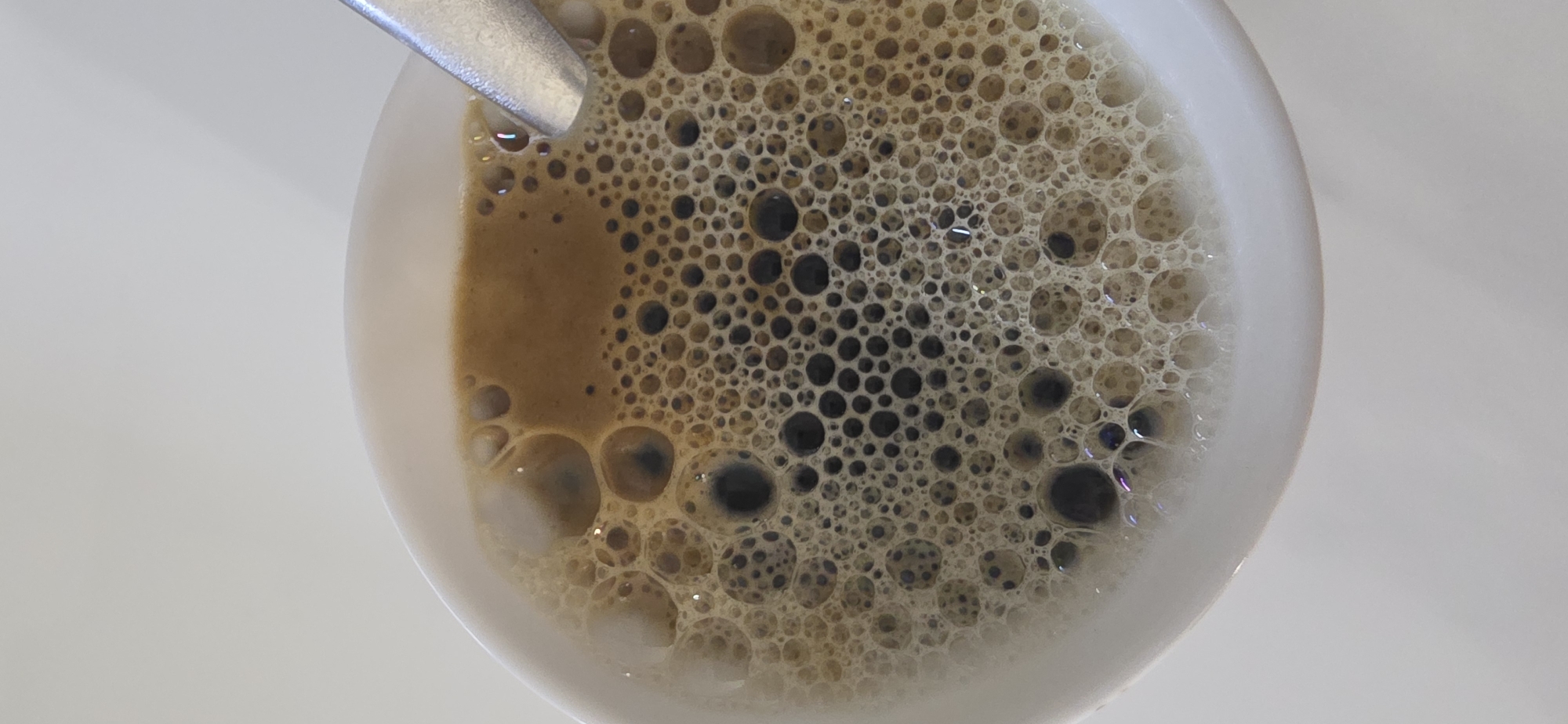 A close-up, overhead shot shows a white cup of coffee with a foamy surface. The coffee is a rich brown color, and the foam is composed of numerous small bubbles, some of which appear darker. A silver spoon rests on the right side of the cup, partially submerged in the coffee. The background is a plain, light-colored surface, suggesting an indoor setting. The image does not contain any specific indicators of location within Sumgayit, Azerbaijan.
