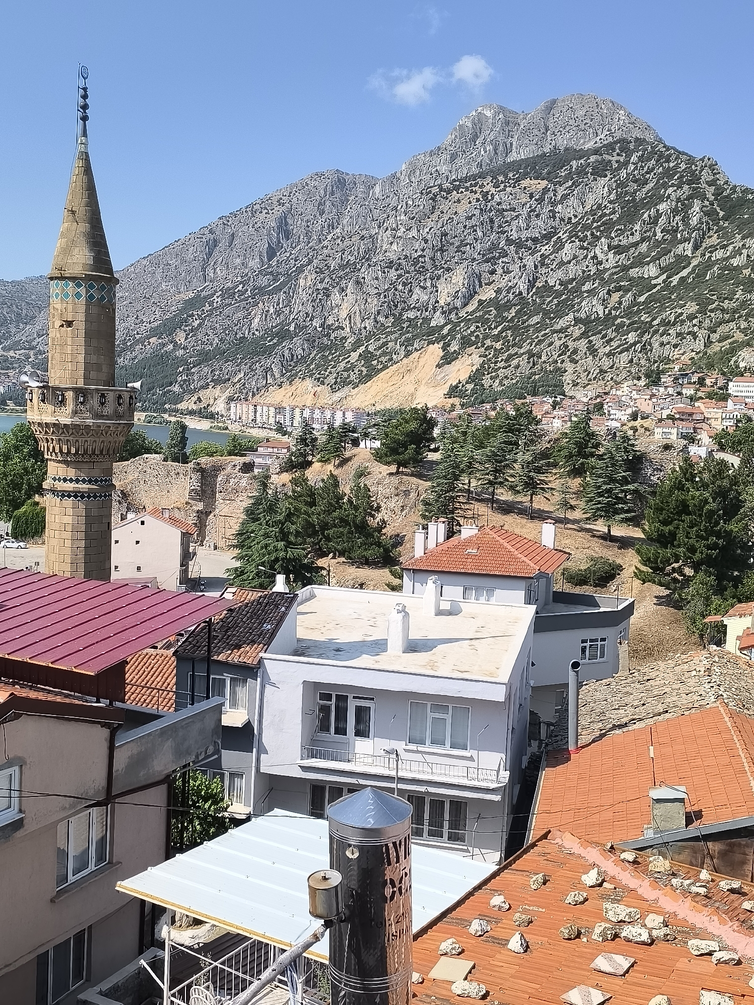 The image captures a view of Eğirdir, Turkey, dominated by a towering minaret in the foreground left. The minaret, constructed from tan-colored stone, features decorative bands of turquoise and green tiles and multiple balconies, each with what appear to be loudspeakers. Behind the minaret, a body of water, likely a lake, is visible, bordered by a town built on a hillside. This town consists of numerous buildings with red-tiled roofs, interspersed with trees and a prominent, rugged mountain range in the background. The foreground of the image is filled with the rooftops of several houses, varying in color and material from red tile to white metal. The overall scene is one of a sun-drenched day, with a clear blue sky above.