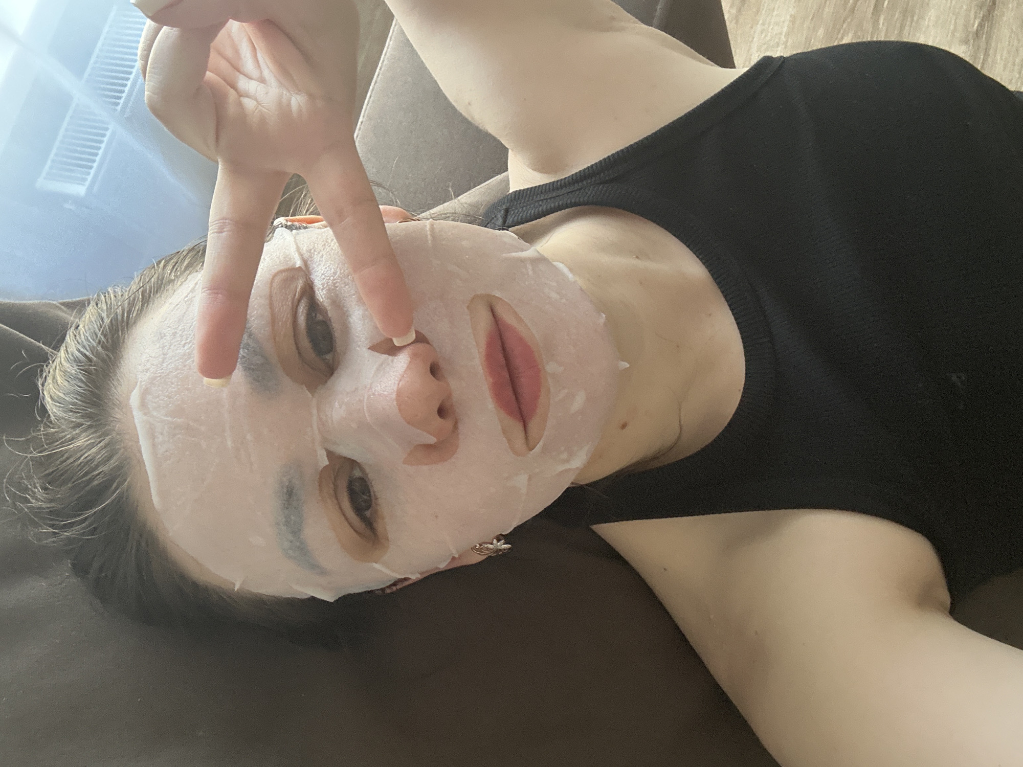 A woman with a facial mask is lying on a brown surface, possibly a couch or bed. She is wearing a black top and making a peace sign with her fingers near her face. A window with a light source is visible in the background. There is no verifiable information that definitively places this image in Chaikovsky, Russia.