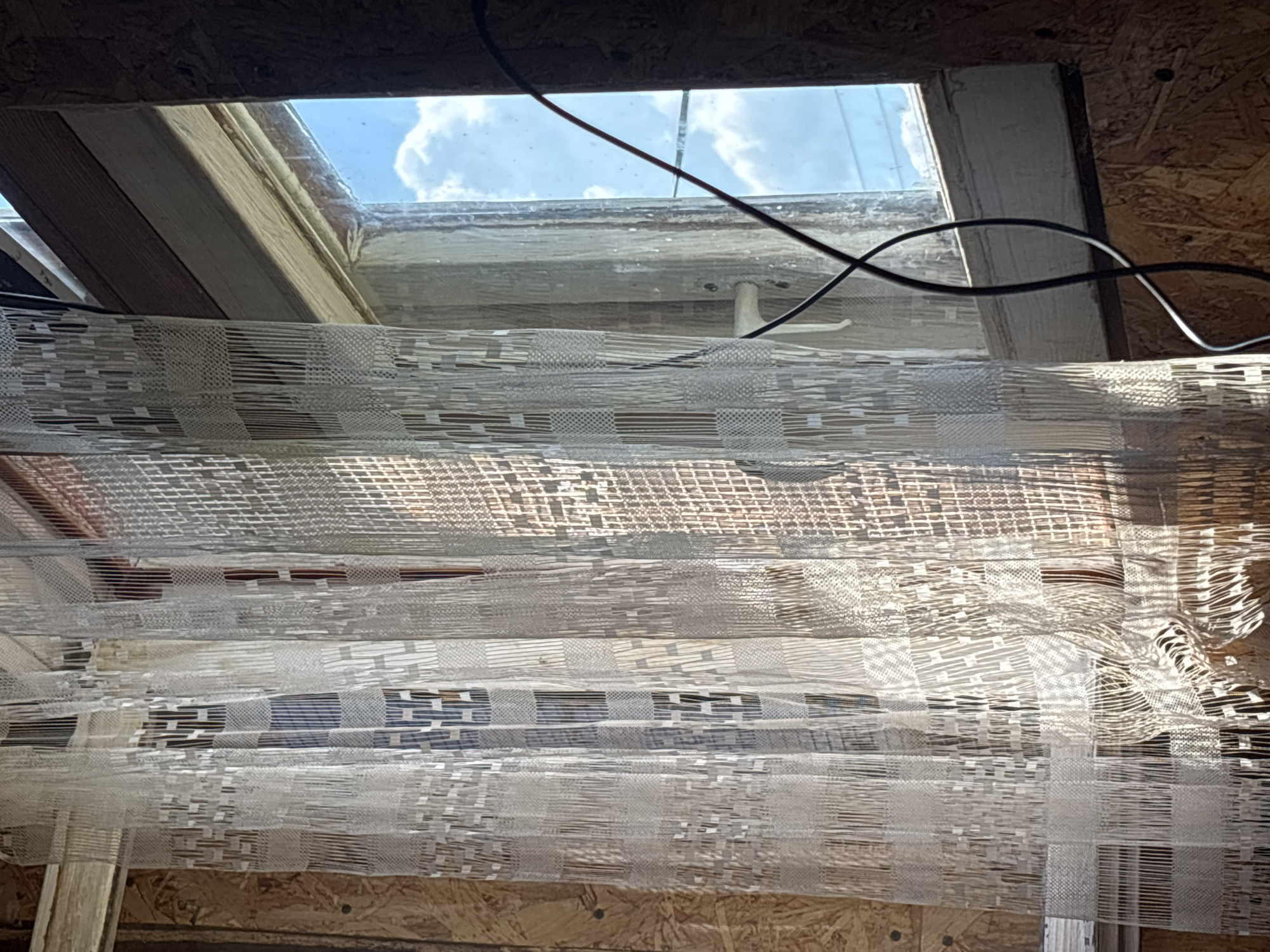 The image captures a view from under a skylight, looking upwards. The skylight frame appears to be made of wood and is set into a ceiling constructed of particle board. The glass of the skylight is somewhat dirty, with a few thin, dark wires running across its surface. Outside, a bright blue sky with fluffy white clouds is visible, suggesting it is daytime.
Dominating the foreground is a series of draped sheer curtains. These curtains have a patterned weave, creating a textured appearance with alternating bands of dense and open fabric, forming geometric shapes. The light filtering through the skylight and the curtains casts shadows and highlights, adding depth to the scene.
The overall impression is one of an unfinished or under-construction interior space. The exposed particle board ceiling and the utilitarian nature of the wiring and skylight frame suggest a functional rather than decorative setting. The presence of the curtains implies an attempt to soften the harshness of the interior or to control the light. There is no visible text in the image. The location context of Yanevo, Russia, does not provide specific visual cues within the image itself.