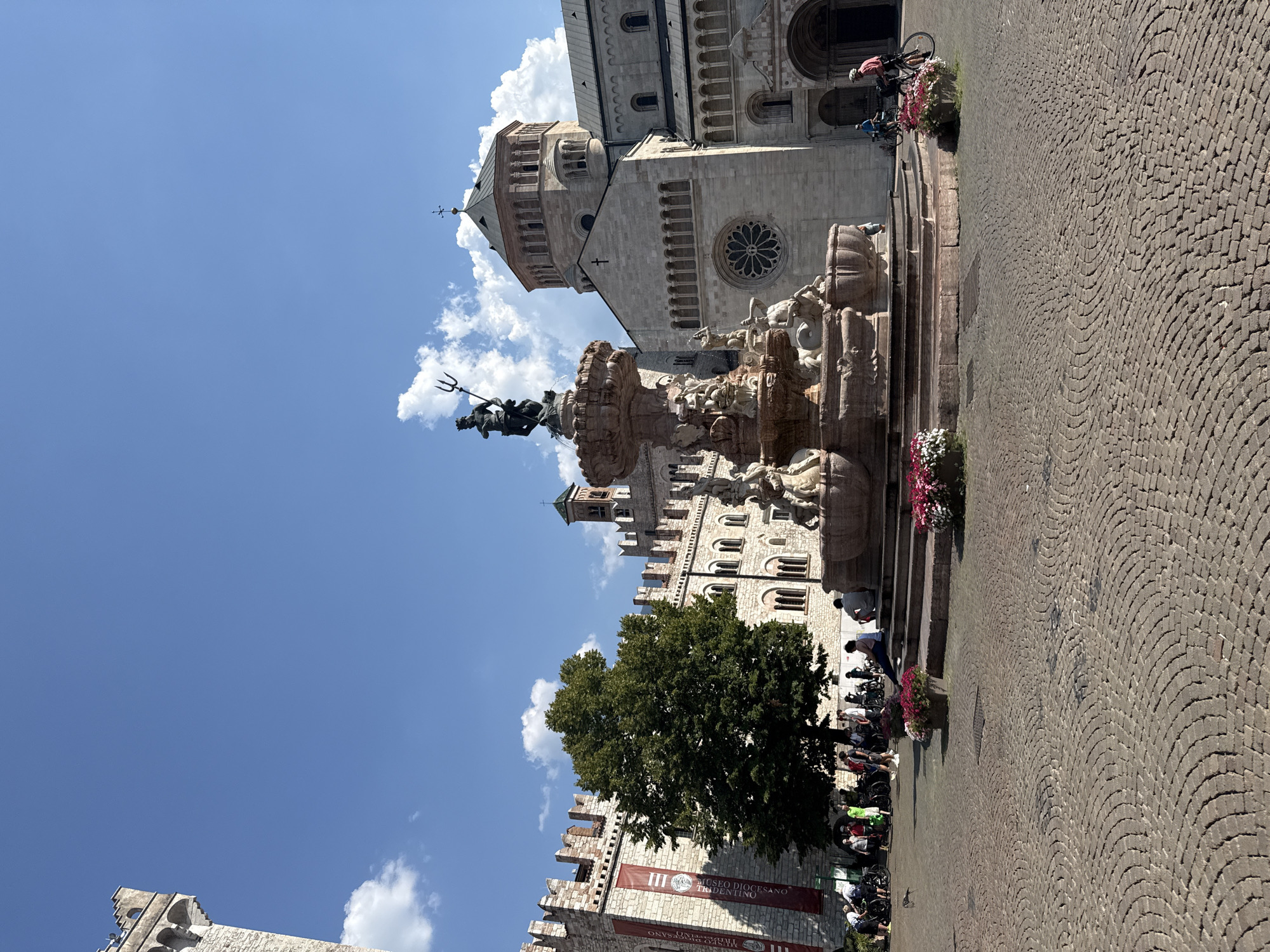 The image depicts Piazza Duomo in Trento, Italy, featuring the Fountain of Neptune and the Trento Cathedral. In the foreground, a cobblestone plaza is partially visible, with potted flowers in shades of pink and white flanking the fountain's steps. The Fountain of Neptune, a large Baroque sculpture, dominates the center of the plaza, with its central figure of Neptune holding a trident.
To the left of the fountain is a section of a stone building with crenellated battlements, likely part of the Palazzo Pretorio or a similar historic structure, adorned with a red banner. A large green tree stands between this building and the fountain.
The Trento Cathedral (Duomo di Trento), a Romanesque-Gothic cathedral, occupies the right side of the frame. Its facade features a large rose window and an entrance archway. The cathedral's prominent dome is visible against a bright blue sky with scattered white clouds.
Several people are present in the scene, some seated on the fountain's steps, others standing or walking around. A group of people with bicycles are gathered near the building on the left, suggesting a popular public space. Two individuals are seen riding bicycles in the middle ground, near the cathedral. The overall atmosphere is one of a sunny, active public square.