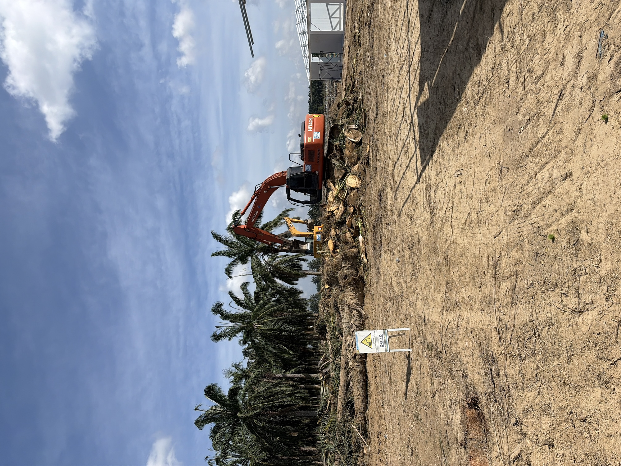 The image depicts a land clearing operation in Simpangempat, Indonesia. An orange Hitachi excavator is positioned amidst felled palm trees and wood debris. A yellow backhoe loader is also present. A warning sign, marked with a triangular hazard symbol and Indonesian text, stands on the cleared ground. In the background, a partially constructed metal-framed building is visible. The sky is blue with scattered clouds. The ground is primarily bare earth, with visible tire tracks and scattered vegetation.