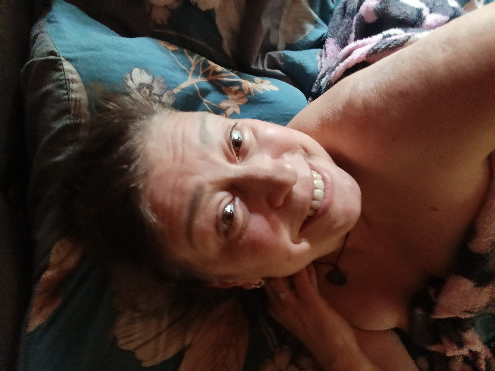 An overhead shot shows a person lying in bed, with their head turned towards the camera and their eyes looking up. The person has their mouth open in a smile, revealing their teeth. They are wearing a necklace. The bedding appears to be a floral pattern with shades of blue and brown. The person is covered by a blanket with a pink and gray pattern. The image is dimly lit, with the person's face being the most illuminated area. The location is not specified in the city of Unknown, Unknown.