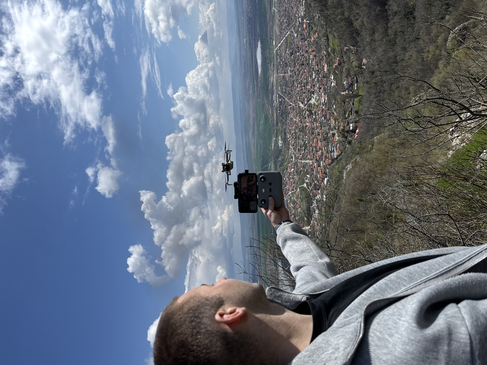 A man is looking up at a drone in the sky. He is holding a controller and a phone in his hand. The background shows a town and a blue sky with clouds. The location is potentially in the city of Shumen, Bulgaria, based on the visible elements. The image captures the man's interaction with drone technology in an outdoor environment.