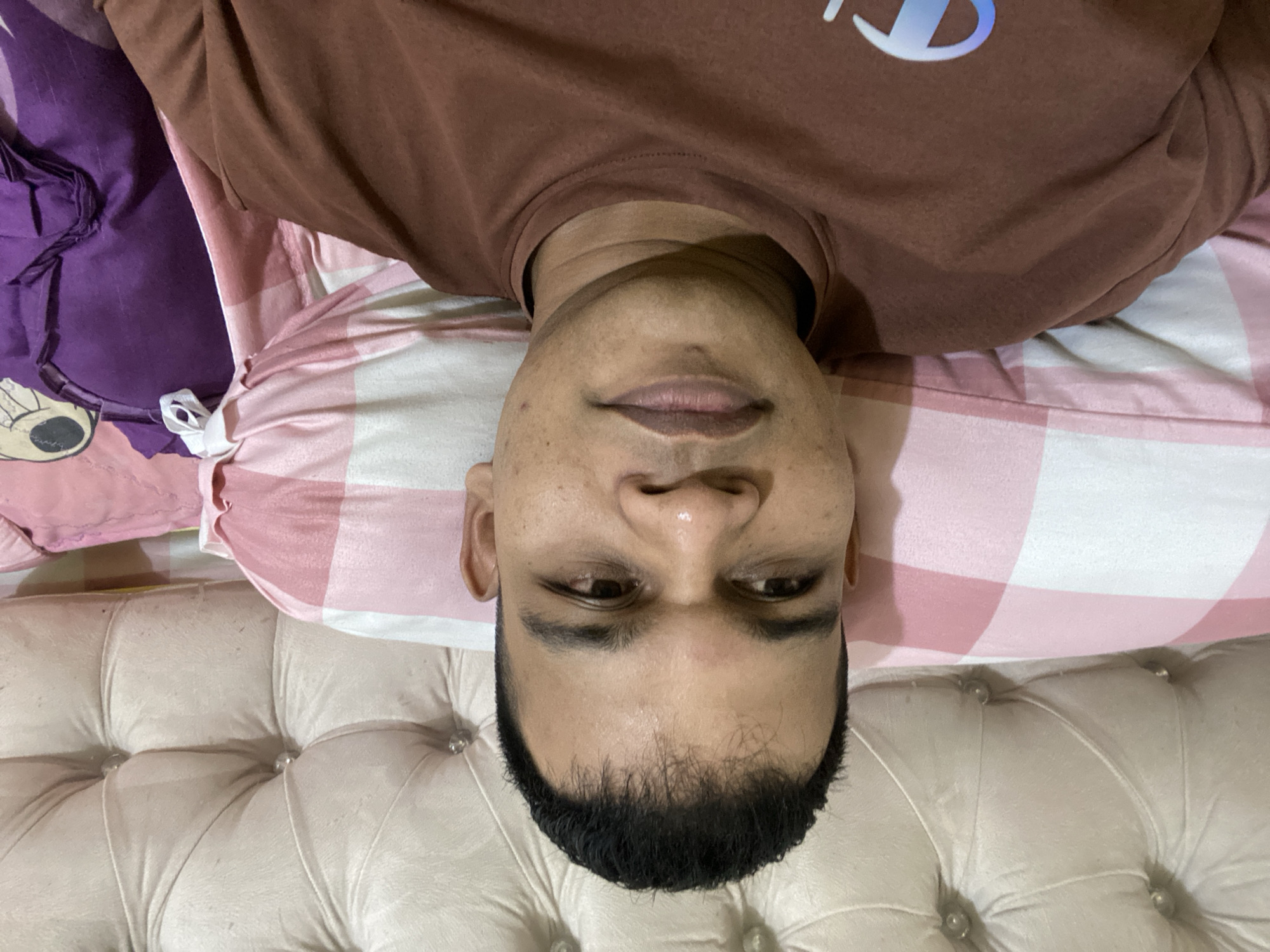 A medium shot shows a man lying down on a bed, facing the camera. He is wearing a brown t-shirt with a white and blue logo on the left chest. The man has dark hair and dark eyes. He is looking slightly to his left. The background consists of a white tufted headboard and pink and white checkered pillows. A purple pillow with a cartoon character design is visible on the right side of the frame. The overall setting appears to be a bedroom in Lambaro Kueh, Indonesia.