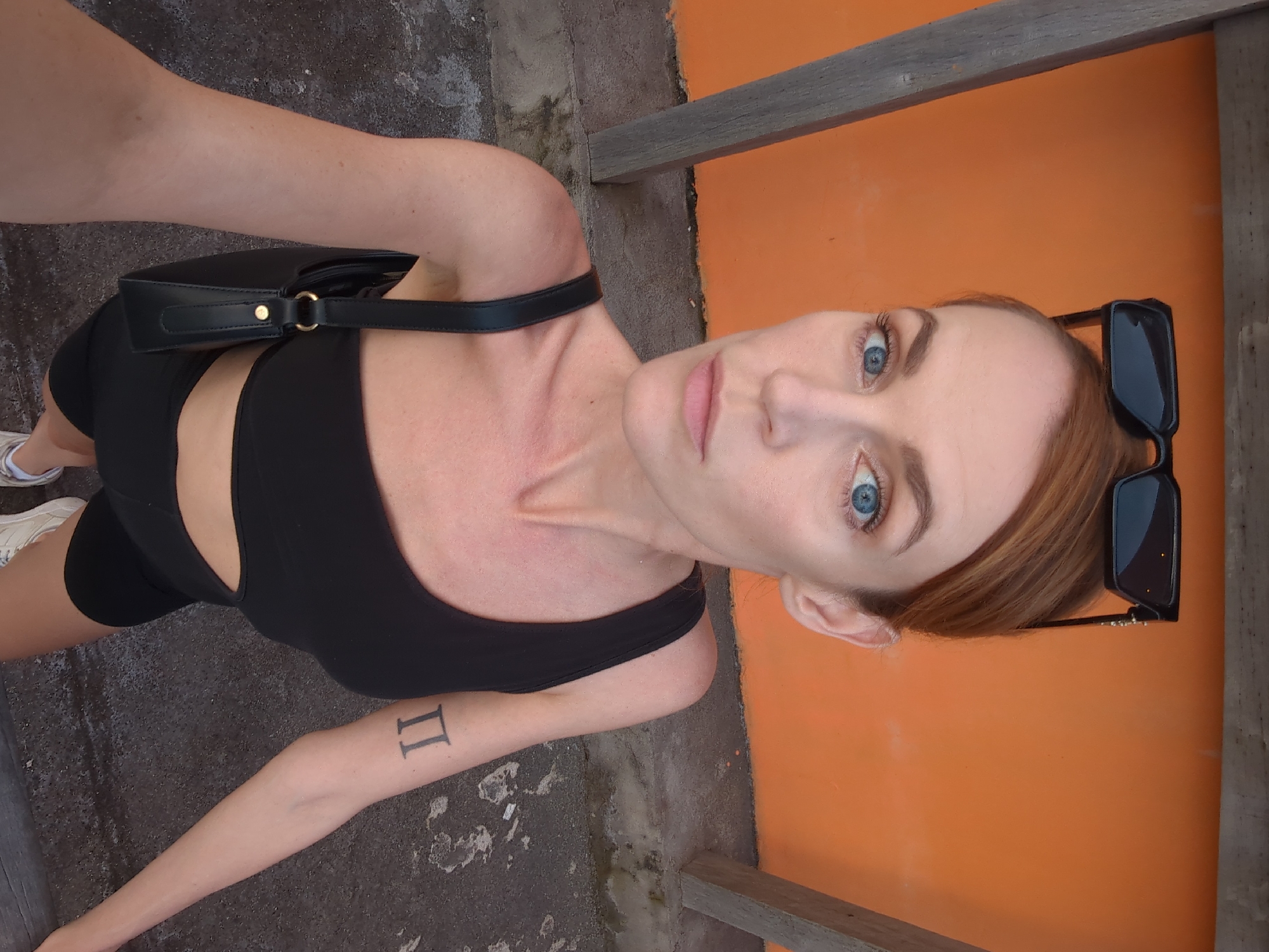 A young woman is captured in a high-angle, tilted selfie, looking directly at the camera with a neutral expression. She has striking blue eyes, light skin, and light brown or reddish hair pulled back, with black sunglasses resting on her head. She is wearing a black crop top or sports bra and matching black athletic shorts, with a black shoulder bag slung over her right shoulder. White sneakers are visible on her left foot. A tattoo depicting the Roman numeral "II" is visible on her left forearm.
The background features a vibrant orange wall on the right side, contrasting with a dark, textured ground, possibly concrete or asphalt, on the left and bottom. Wooden beams or structural elements are visible at the top and bottom edges, suggesting an outdoor setting like a balcony or walkway. The bright lighting indicates it is daytime. The scene is likely set outdoors in North Kuta, Indonesia, reflecting a warm climate.