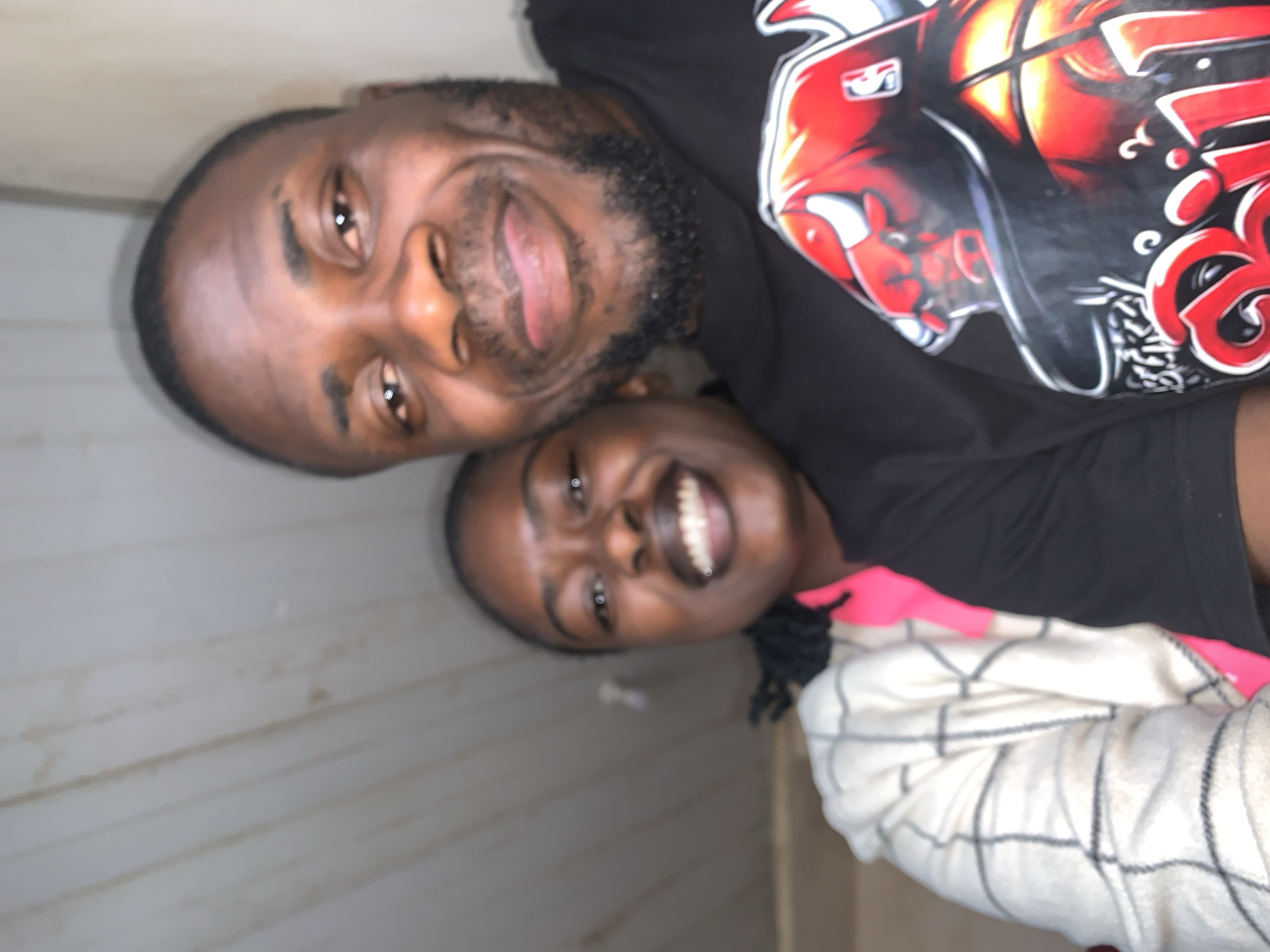 Two adults, one male and one female, are depicted in a close-up, chest-up shot. The male, positioned in the foreground on the right, has a dark complexion, a short beard, and short hair. He is smiling broadly, revealing his teeth, and wears a black short-sleeved t-shirt with a large red and black graphic featuring a bull's head (Chicago Bulls logo), a basketball, and the word "Bulls." The female, partially obscured behind and to the left of the male, also has a dark complexion and dark hair. She is smiling broadly, revealing her teeth, and wears a light-colored (possibly cream or off-white) jacket or cardigan with a dark grid pattern. A bright pink garment is visible underneath her jacket. Both individuals are looking directly at the camera, suggesting a posed photograph. The background is an indoor setting with a light-colored ceiling or upper wall composed of vertical panels or planks, showing some faint lines and minor discoloration. A small, indistinct light source is visible in the upper background. The scene is situated in the city of New Stadium, Nigeria.