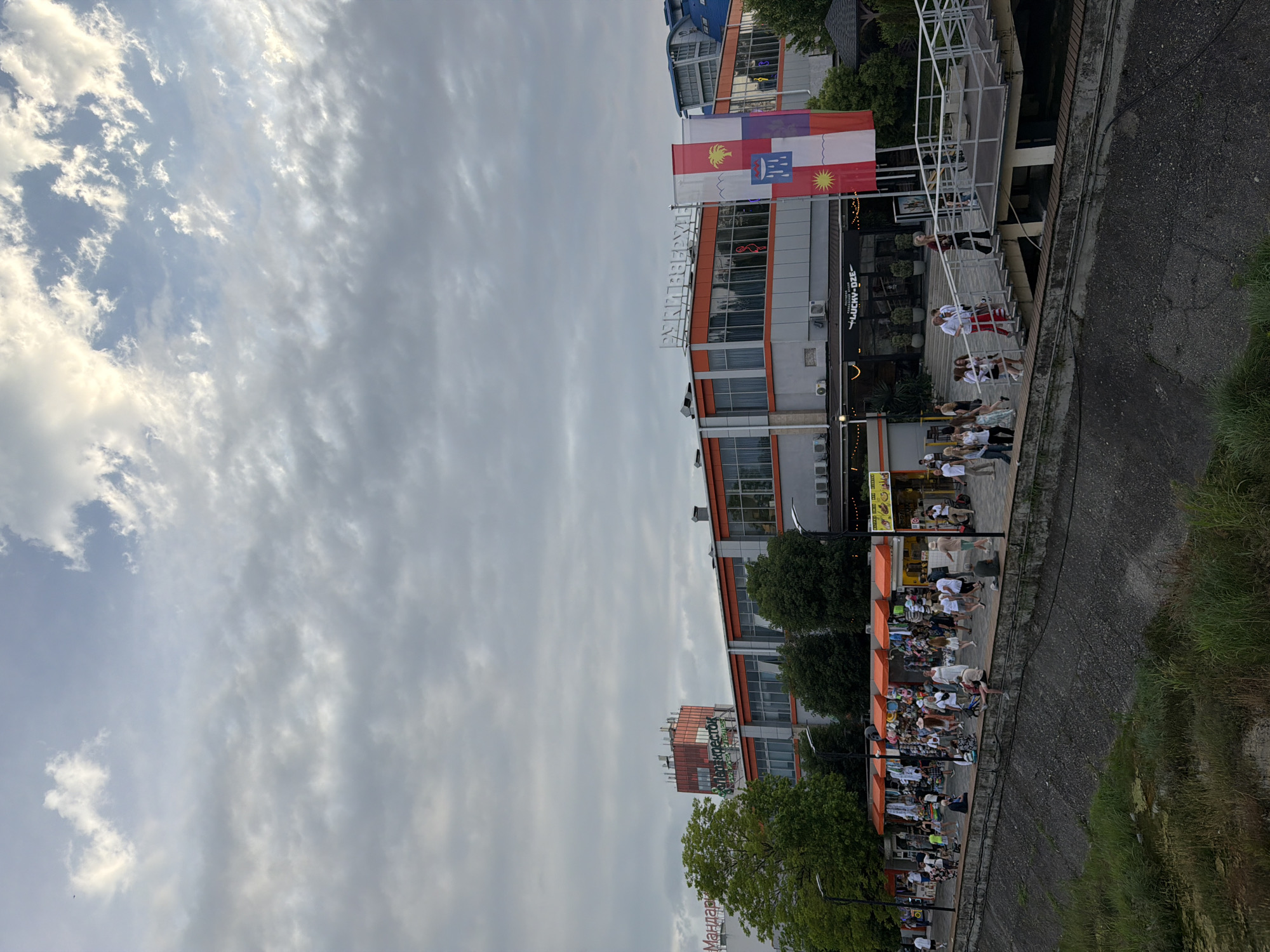 The image shows a waterfront promenade in Sochi, Russia, likely near the Sochi Maritime Station or a similar area featuring shops and restaurants. The sky is overcast with dramatic clouds. The promenade is lined with several buildings, including a multi-story structure with orange trim and large windows, and a prominent orange building topped with a red structure that has Cyrillic text, likely a business or landmark. Flags are displayed in front of one of the buildings.
Numerous people are walking along the promenade, some browsing stalls with orange awnings, others conversing or walking towards a ramp leading to a higher level. The area appears to be a popular spot for leisure and shopping. The lower part of the image shows a concrete embankment with grass and water. The overall scene depicts a lively urban waterfront environment under a cloudy sky.