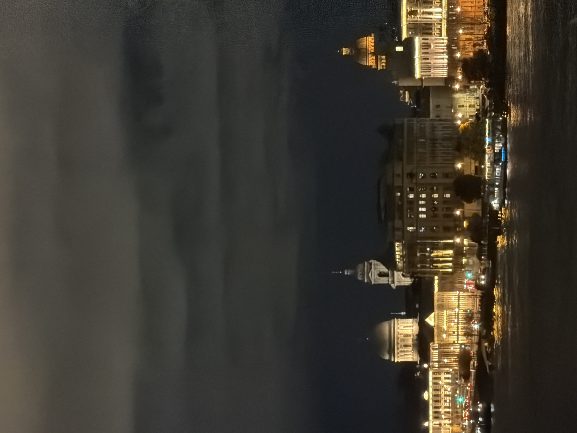 A nighttime cityscape view of Saint Petersburg, Russia, showcases illuminated buildings along the Neva River. The buildings are characterized by ornate architecture, with domes and multiple stories, all lit from within and by external lighting. Lights are also reflected on the dark water of the river, suggesting movement and depth. Several vehicles are visible on the road in front of the buildings, and a few boats are present on the river. The sky above is dark and cloudy, with visible cloud formations. The overall impression is of a vibrant, well-lit urban environment at night.