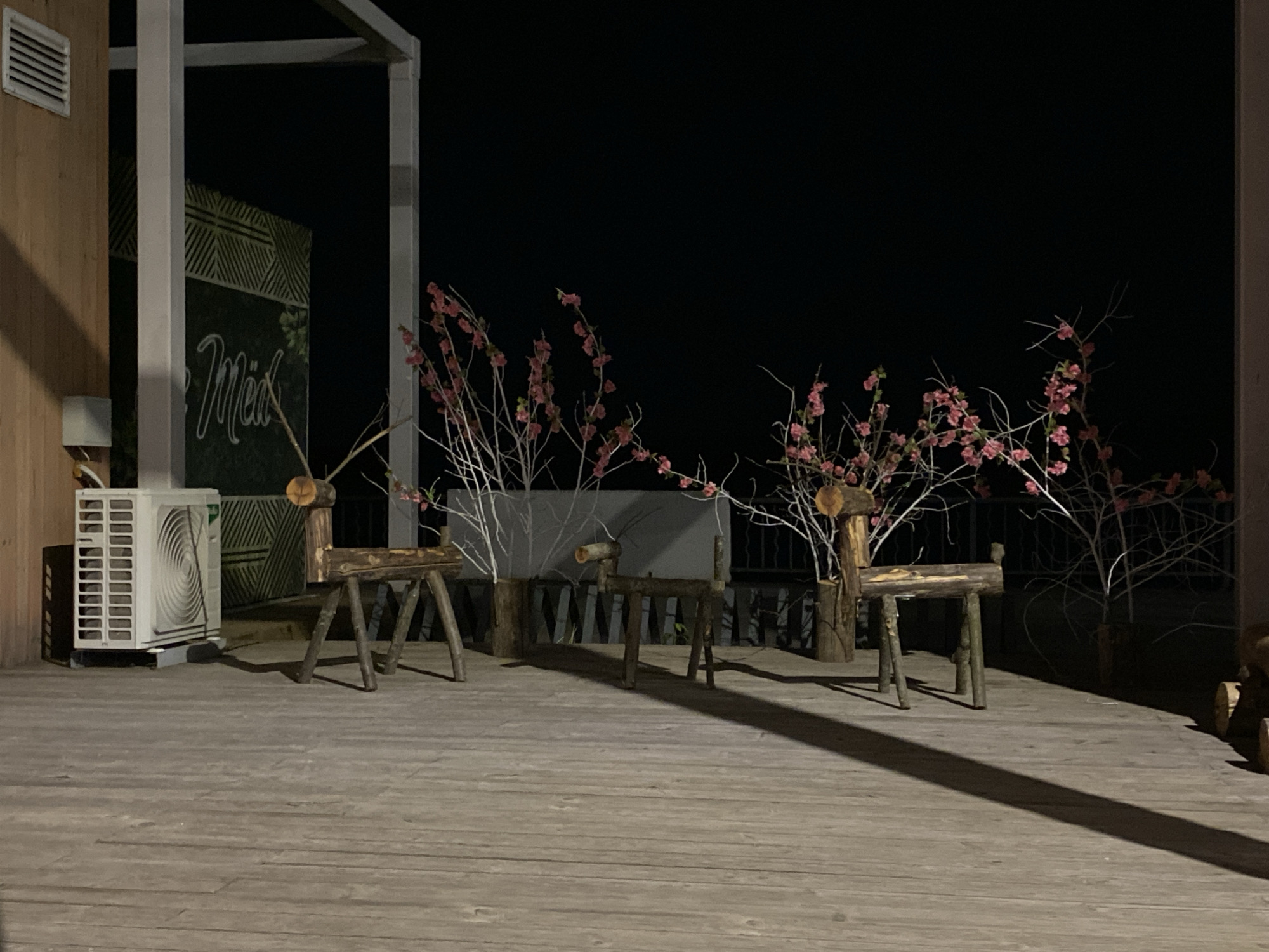 The image shows a dimly lit outdoor patio in what appears to be the city of Chaikovsky, Russia, at night. The patio has a wooden floor and is partly covered by a wooden structure. A sign with Cyrillic script (likely "Мёд") is visible. An air conditioning unit is located near the corner of the building. Several handmade wooden reindeer statues are arranged along the edge of the patio, interspersed with white branches adorned with small pink flowers. The background is dark, suggesting a nighttime scene.