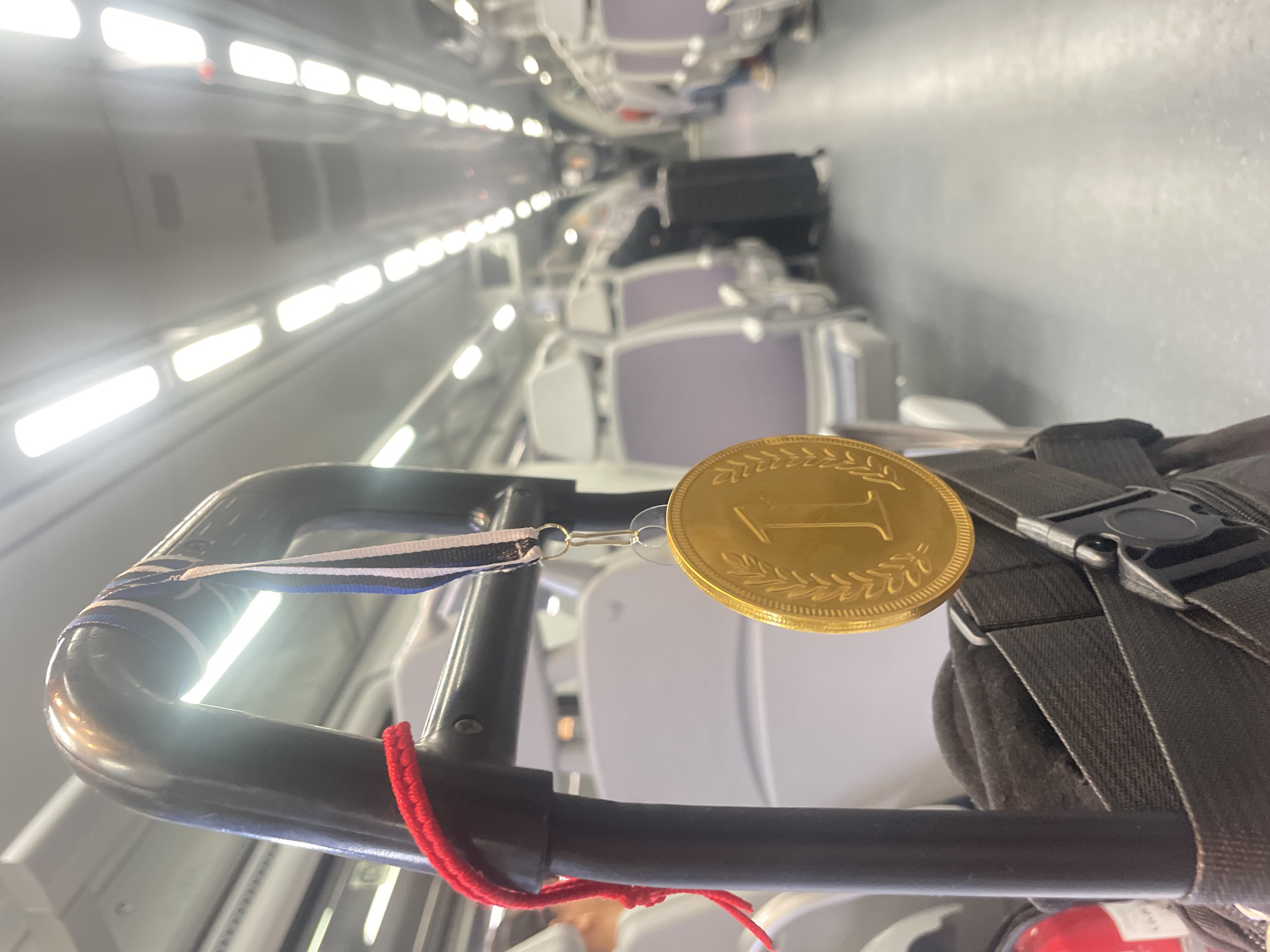 The image shows the interior of a train car, likely traveling through Barcelona, Spain. The perspective is looking down the aisle, with rows of passenger seats on either side. Overhead lights illuminate the scene. Attached to a luggage handle is a gold medal with the number "1" on it. There is a white, blue, and black ribbon attached to the medal, and a red string is tied to the luggage handle. A grey backpack is also attached to the luggage handle.