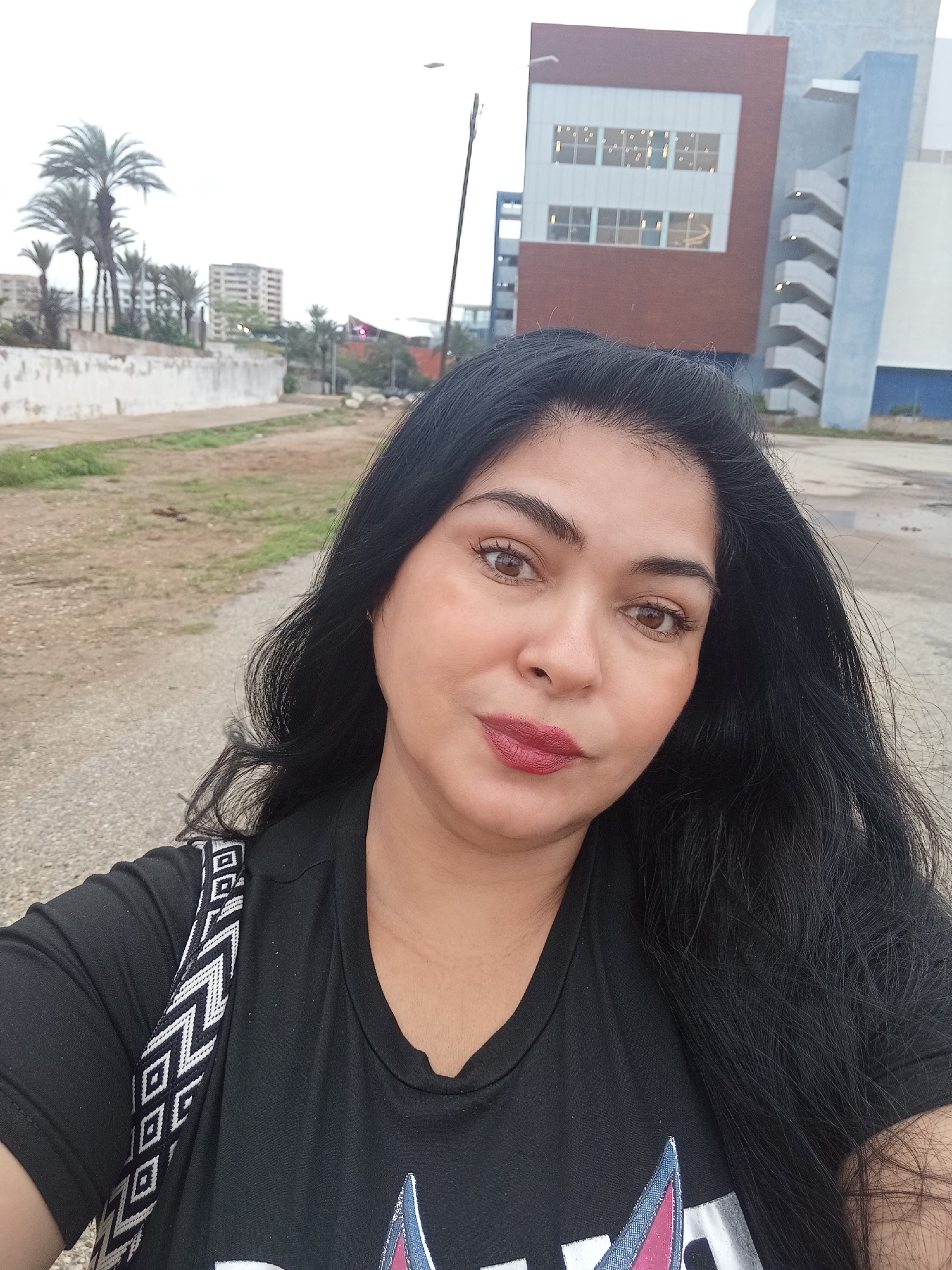 A medium shot captured from a low angle shows a woman with long, dark hair in the foreground, facing the camera. She is wearing a black t-shirt with a patterned strap of a bag visible over her left shoulder. Her makeup includes dark eyebrows and red lipstick. Her eyes are brown and she has a slight smile.
The background is an outdoor urban setting. To the left, there is a low, white wall with some vegetation and palm trees behind it. Further in the distance, several buildings are visible, including a tall, light-colored building. To the right of the frame, a modern building with a red and white facade, featuring multiple windows, stands out. A blue and light grey structure with an external staircase is attached to it. The ground is a dirt and gravel surface, with some puddles of water suggesting recent rain. The sky is overcast.