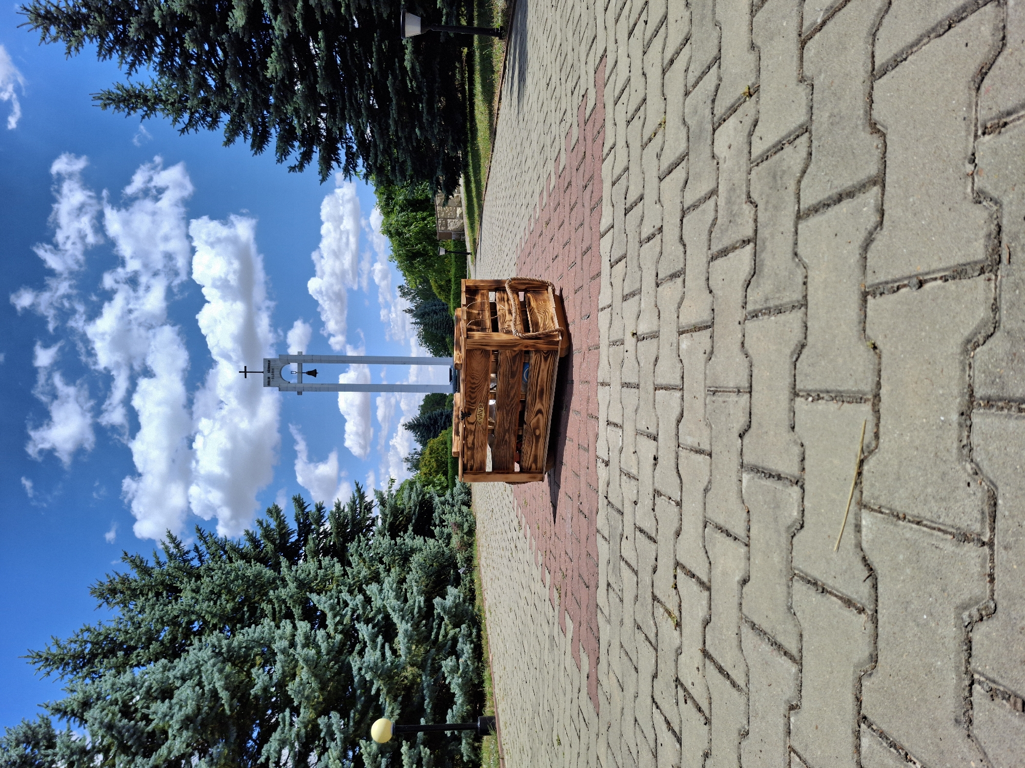 A wooden crate is positioned centrally on a paved walkway in a public outdoor space. The walkway is constructed of interlocking concrete pavers in a grey and red pattern. The crate, made of burnt wood, is placed directly in front of a stone memorial or monument with a tall, narrow structure topped by a cross and a bell. Flanking the walkway are rows of tall evergreen trees. In the background, the sky is bright blue with scattered white cumulus clouds. Two lampposts are visible, one to the left and one to the right of the walkway, partially obscured by the trees. The scene is set during daylight hours under a clear sky. The location is identified as Șerpeni, Moldova. There are no people or animals present in the image.