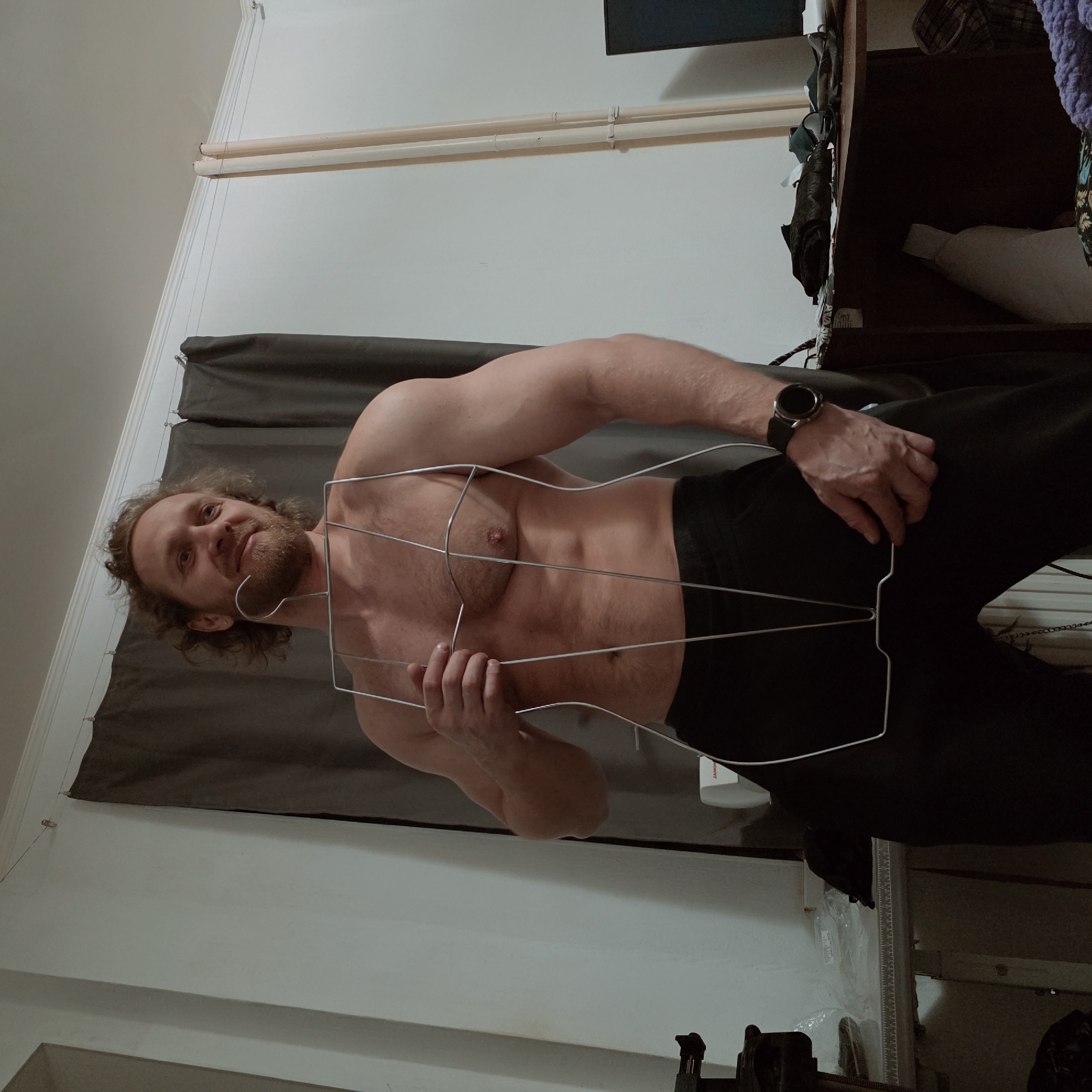 A male individual, appearing in his 30s to 40s with medium-length wavy hair and a beard, stands bare-chested, wearing black pants and a dark smartwatch on his left wrist. He is holding a metallic wireframe sculpture shaped like a human torso in front of his body, with his right hand grasping the upper-left side and his left hand near the lower edge. He displays a slight smile and looks towards the viewer's left.
The scene is an indoor setting, likely a room with light-colored walls and white crown molding near the ceiling. Dark grey curtains hang from a thin wire stretched across the upper wall, secured by clips. To the left, a light-colored table features a ruler marking along its edge, with a white sewing machine (brand "JANOME" partially visible) and a clear plastic bag containing items. To the right, two light-colored vertical pipes run along the wall. Further right, a dark desk or table is visible, cluttered with various items, including fabric, papers, and a dark electronic screen on the far right edge. The specific location within Saint Petersburg, Russia, cannot be determined from the visual information provided.