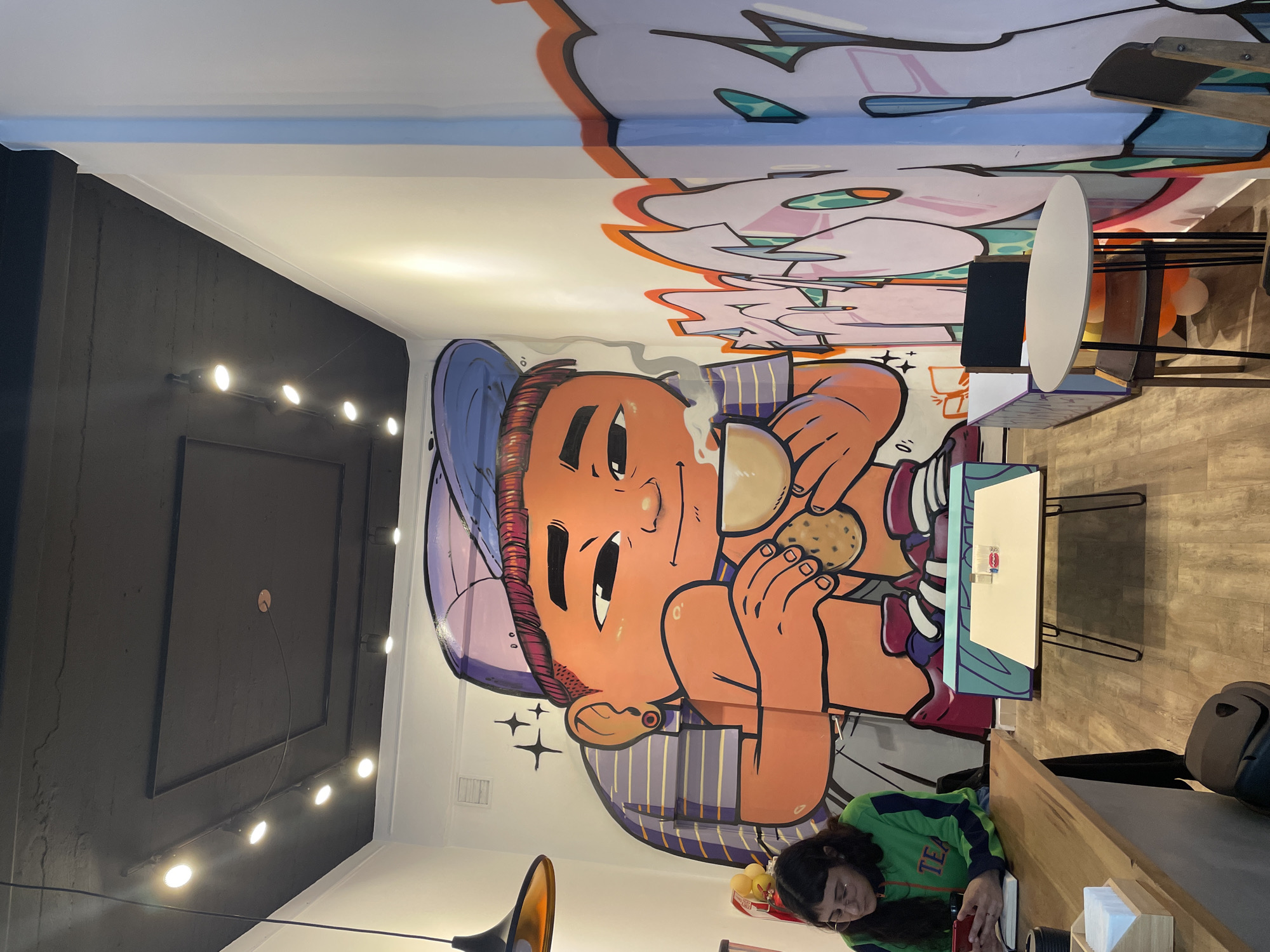 The image captures the vibrant interior of what appears to be a cafe or casual eatery, dominated by a large, colorful mural. The mural features a cartoon-style character, resembling a boy with an orange face, a blue baseball cap, and striped sleeves. He is depicted happily holding a steaming cup in one hand and a chocolate chip cookie in the other. The mural's art style is reminiscent of graffiti, with bold outlines and a playful aesthetic, extending onto the ceiling with abstract shapes and colors.
In the lower foreground, a young woman with dark hair is seated at a wooden counter or table. She is wearing a green and blue jacket with the word "TEAM" visible on the sleeve and is looking down, seemingly focused on an item in her hands, possibly a phone or book.
The room features contrasting elements in its design. One section of the wall or ceiling is dark grey/black, equipped with a linear track lighting system holding several bright, circular spotlights. The floor appears to be made of light-colored wooden planks. Other furnishings include white, oval-shaped tables with thin metal legs and chairs. A lamp with an orange interior hangs in the foreground, and on the counter, there's a wooden holder with tissues and a small basket containing several yellow, spherical objects. The artificial lighting suggests an indoor setting where the time of day is not explicitly discernible.
