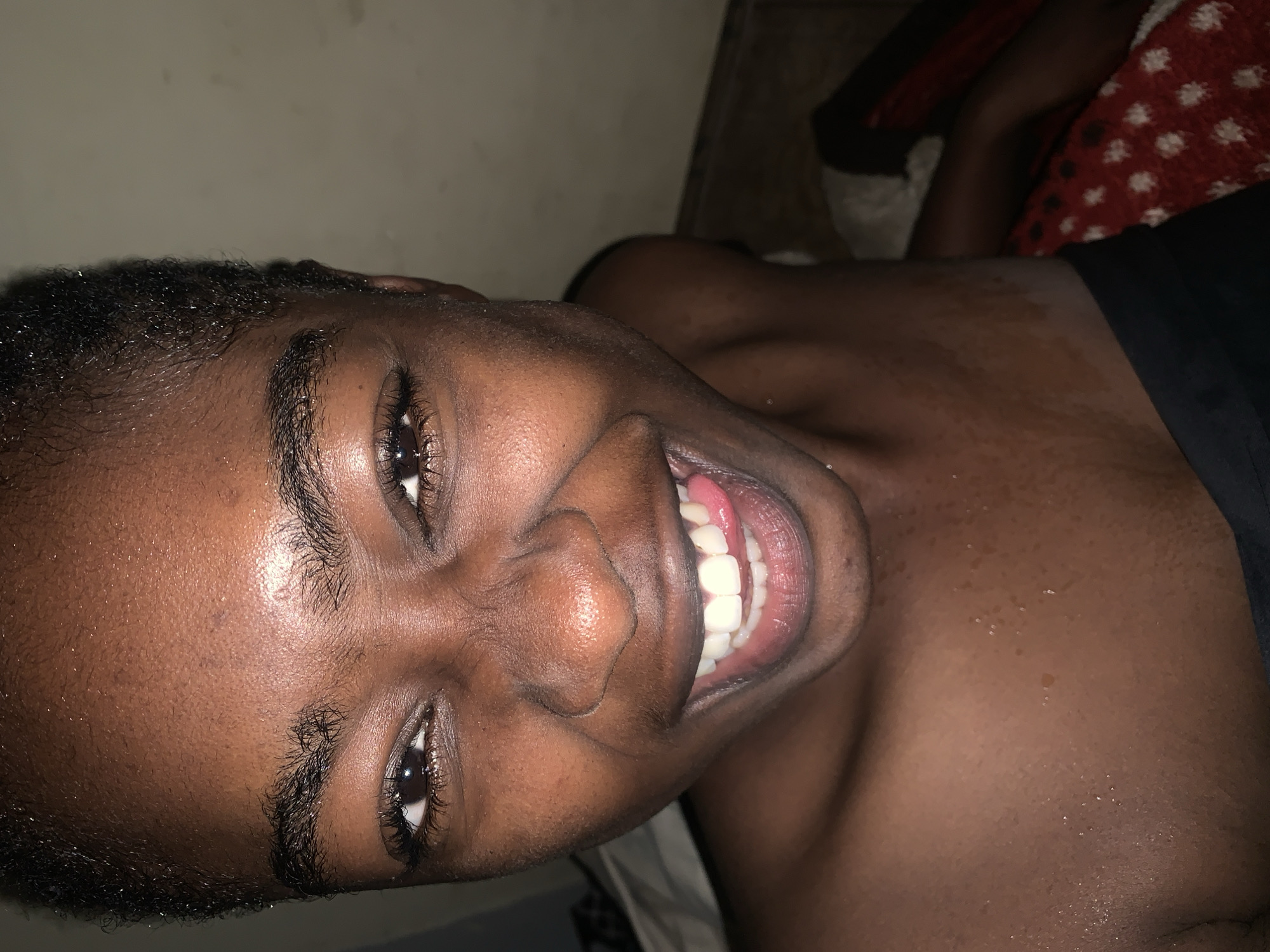 A close-up, head-and-shoulders shot captures a young Black individual with dark skin, short hair, and a radiant smile in Rafin Par, Nigeria. The individual is looking directly at the camera, with their tongue slightly extended, revealing bright white teeth. Their skin appears moist, possibly from sweat or recent bathing. The background is indistinct, with a light-colored wall and a dark floor, suggesting an indoor setting. The lighting is somewhat dim, creating a warm and intimate atmosphere. The individual's facial expression conveys joy and playfulness.