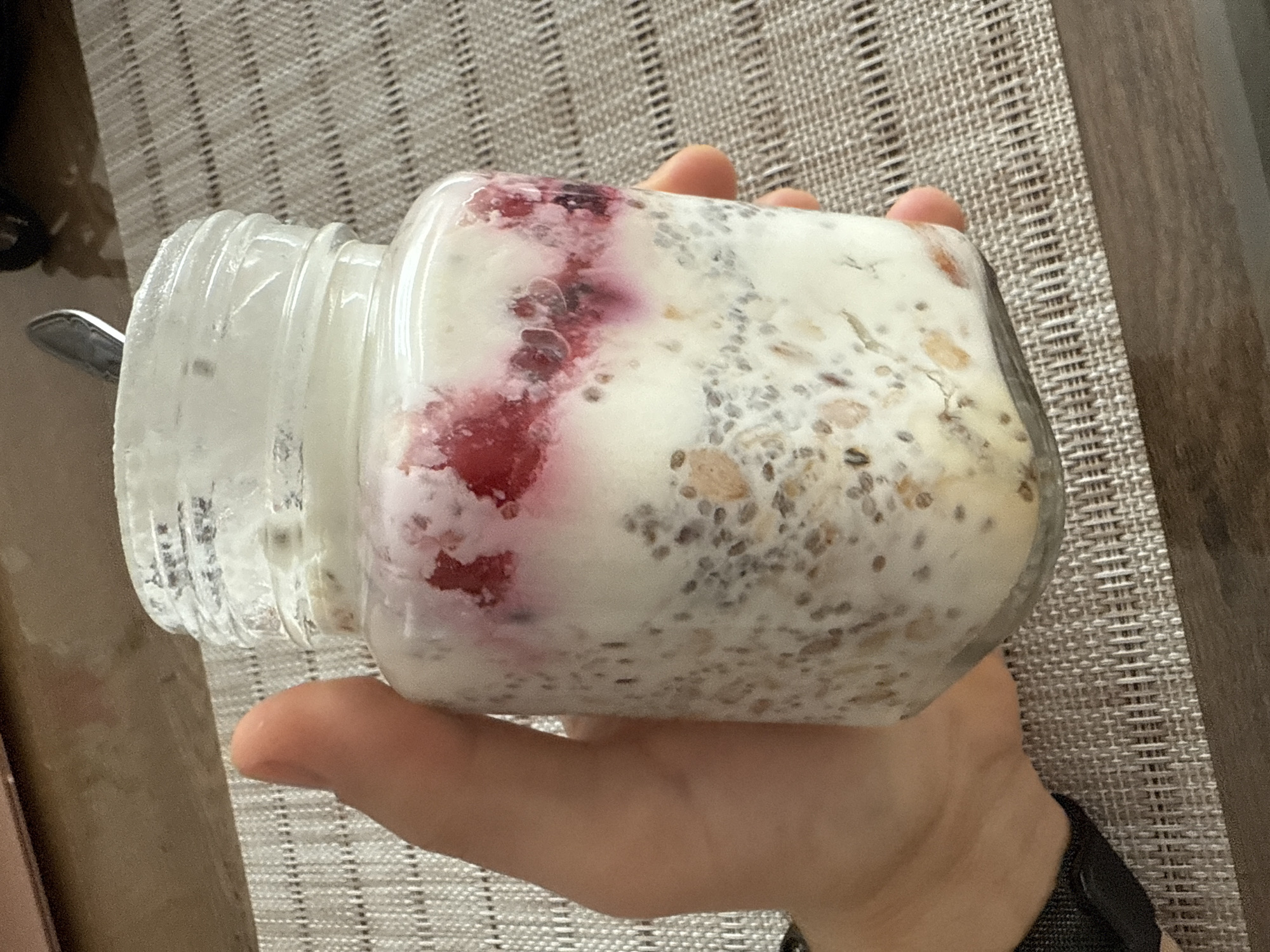A hand holds a jar filled with what appears to be chia seed pudding with red berries. The jar is glass, and a spoon is visible inside. A woven mat is visible in the background, with a wooden table to the right. The image does not contain any information that can be used to infer the location of the scene, including if it is within the city of Sofia, Bulgaria.