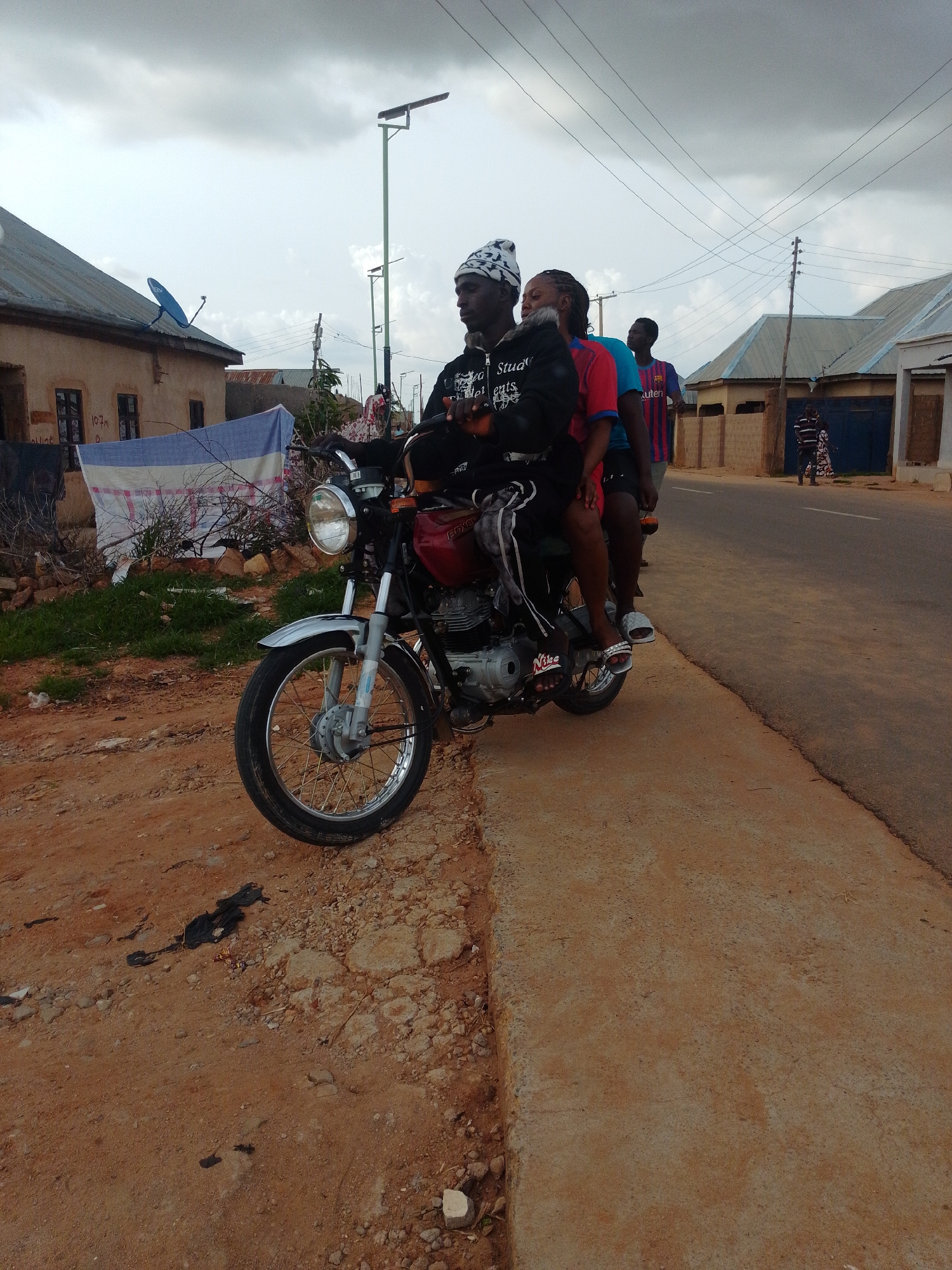 The image depicts a street scene in Gombe, Nigeria, under a cloudy sky. In the foreground, a motorcycle is parked on a dirt and gravel roadside. The motorcycle is red and silver, with a chrome front fender and a spoked wheel. A man wearing a black jacket with white text, a head wrap, and dark pants is seated on the motorcycle. A woman with dark braided hair and a black jacket is seated behind him. Another person, a man wearing a blue and red striped t-shirt, is visible behind them, also on the motorcycle. In the background, there are buildings with corrugated metal roofs, a satellite dish on one of the buildings, and utility poles with power lines. Further down the street, two people are walking. The overall environment appears to be a residential area with a mix of dirt and paved surfaces.