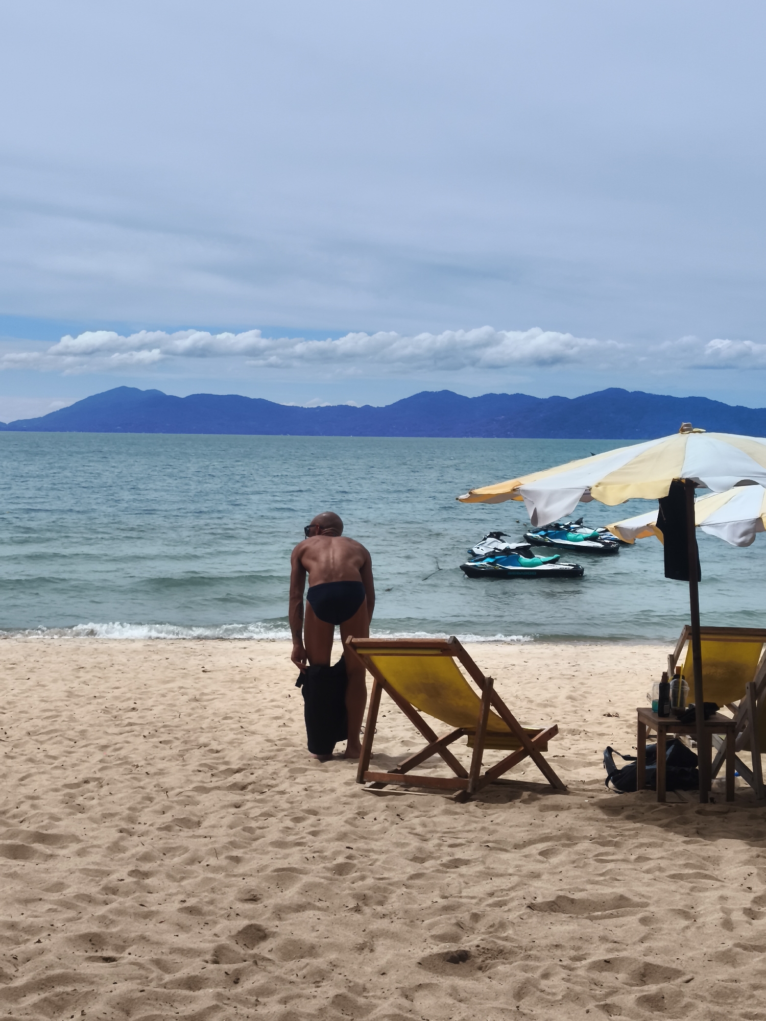 A man wearing black swim trunks is on a sandy beach in Baan Mae Nam, Thailand, bending over to pick up a black bag. The sea is calm with gentle waves, and in the distance, mountains are visible under a cloudy sky. Several jet skis are anchored in the water. Two yellow lounge chairs are set up on the sand, one with a small wooden table beside it. A yellow and white striped umbrella provides shade over the lounge chairs.