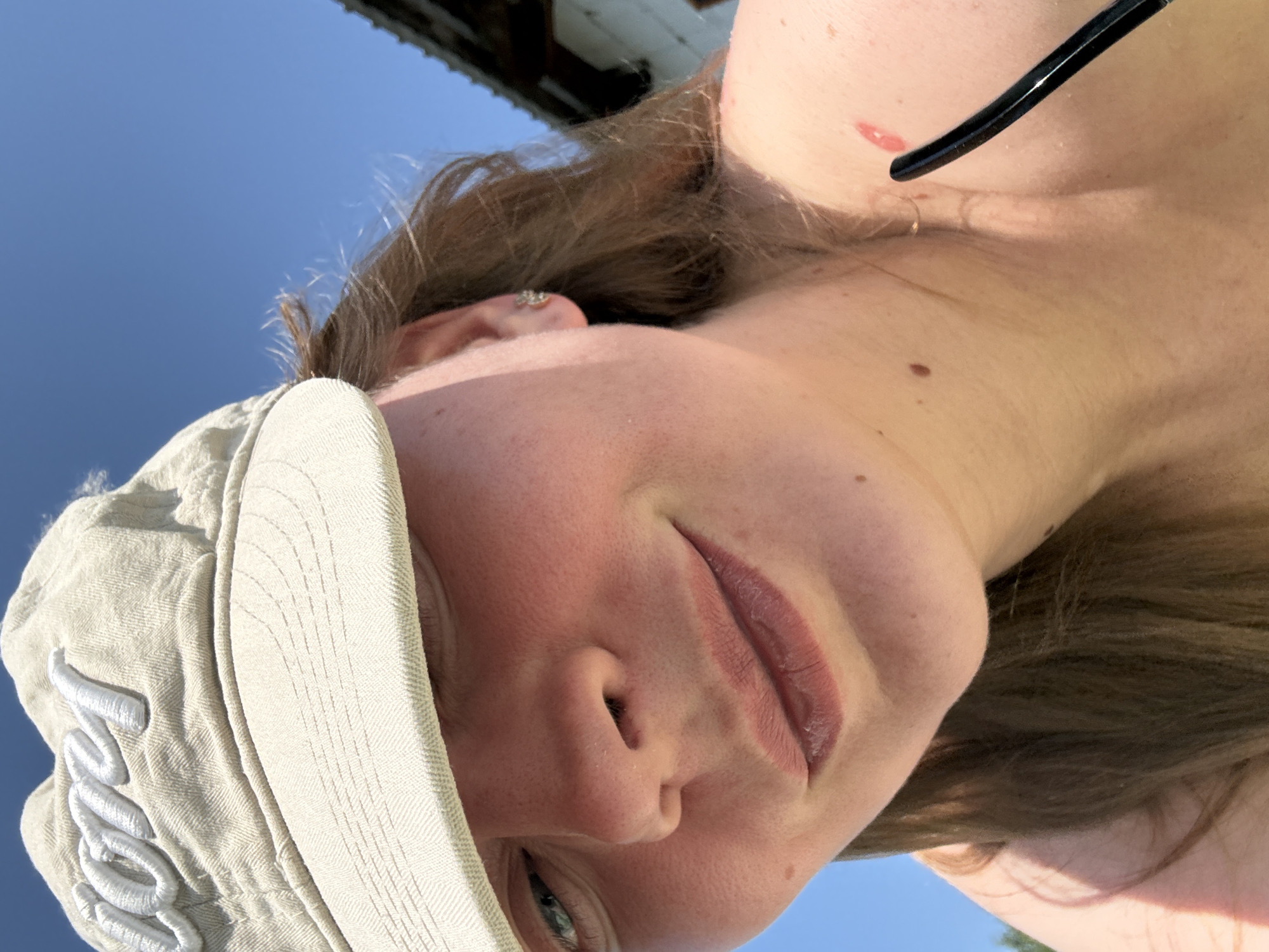 A close-up, low-angle selfie of a fair-skinned person with light brown hair, wearing a light-colored baseball cap. The cap has white embroidered lettering. The person is looking downward, with a visible blue sky in the background. Part of what appears to be a structure is visible in the upper-left corner. The person is wearing earrings and what appear to be sunglasses. The location cannot be determined from the given image.