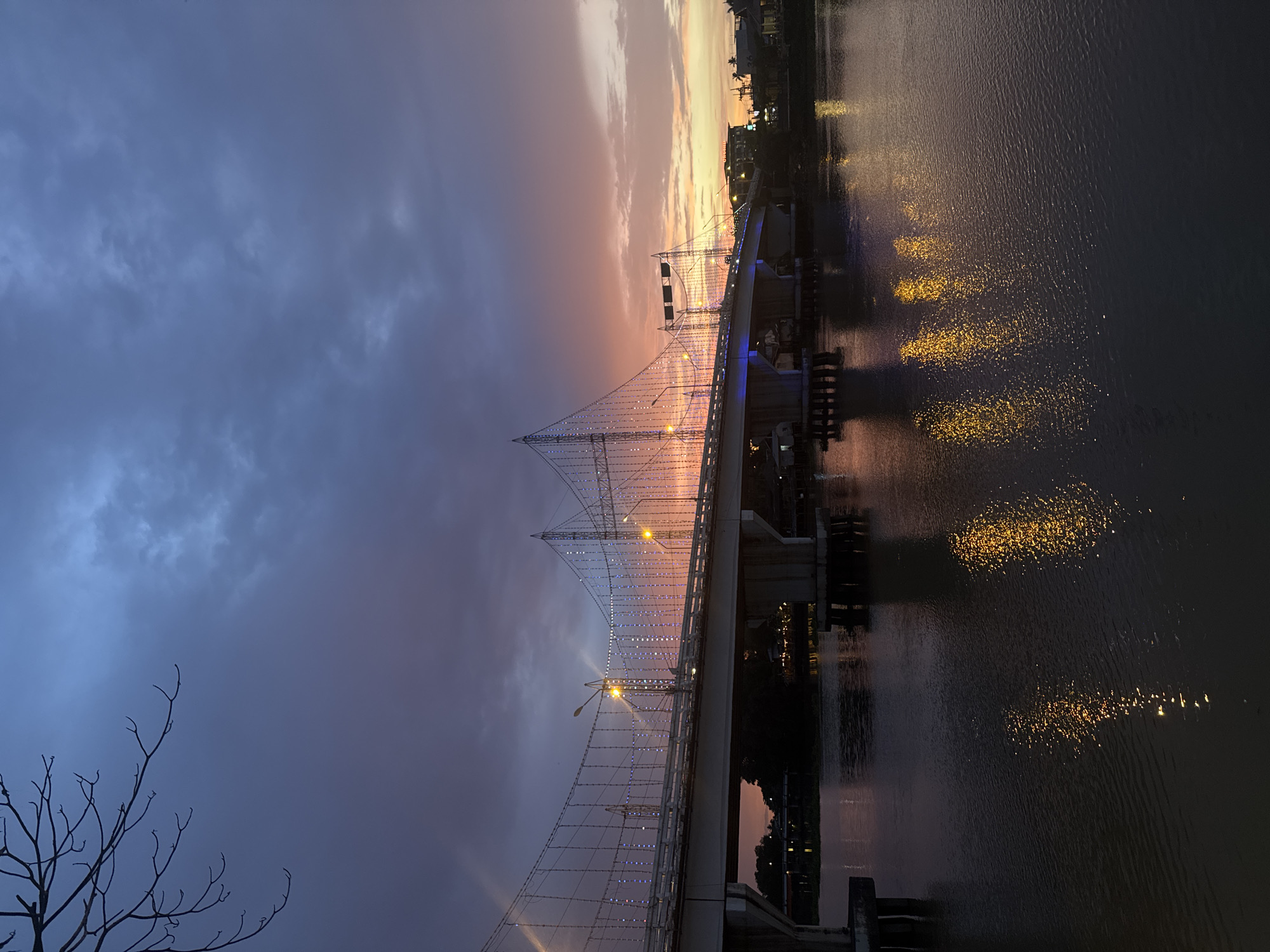 This is a vertical, landscape-oriented photograph of a cable-stayed bridge illuminated with lights over a body of water at dusk. The sky is a dramatic mix of deep blues and grays on the left, transitioning to soft oranges, pinks, and yellows towards the right horizon, indicating sunset. Wispy clouds are visible across the sky. The bridge itself is a prominent feature, adorned with strings of small, white and blue lights along its cables and structure, creating a festive appearance. Yellow lights from the bridge and distant structures reflect on the rippling surface of the water, creating shimmering patterns.
The scene captures a serene yet lively atmosphere as the day transitions into night. The bridge appears to be a significant piece of infrastructure, possibly a landmark in Surat Thani City Municipality, Thailand, as suggested by the location context. There are hints of buildings and palm trees on the far bank of the water, further placing the scene in a tropical or subtropical region. In the lower left corner, the silhouetted branches of a bare tree add a touch of natural contrast to the man-made structures. The overall mood is peaceful and visually appealing, highlighting the beauty of the bridge and its surroundings at twilight. No discernible text is visible in the image.