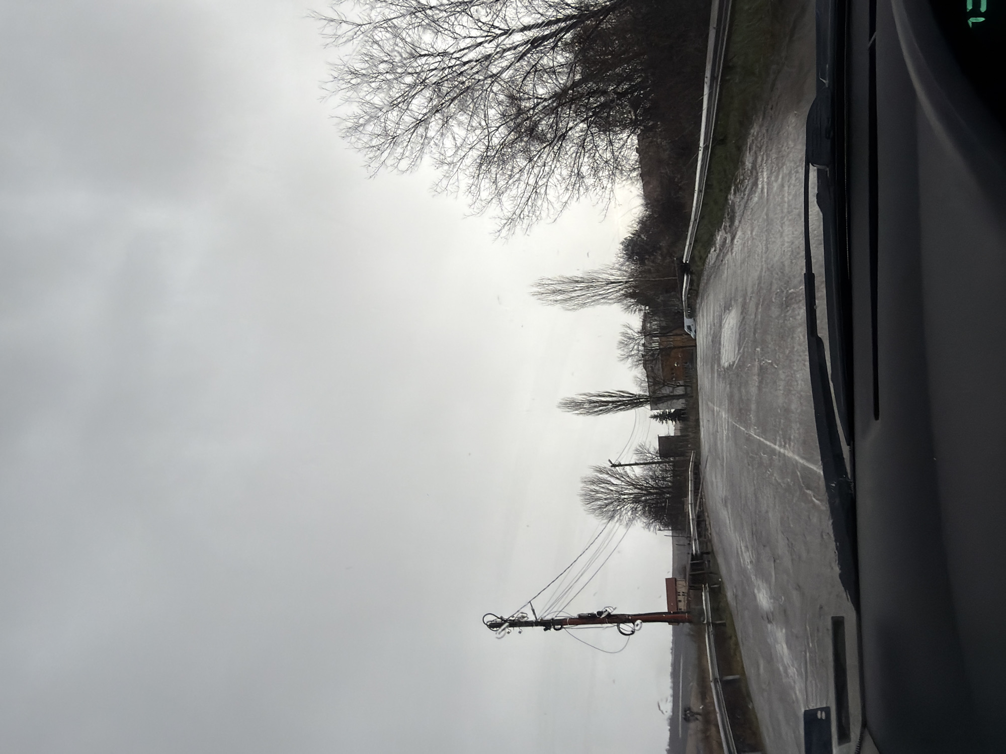 The image presents a view from inside a vehicle on a wet road in Dragichevo, Bulgaria. The sky is overcast, indicative of rain or a recent rainfall. The road stretches ahead with faded white lane markings. Buildings and leafless trees line the side of the road. A utility pole with exposed wiring is visible. A guardrail runs along part of the road.