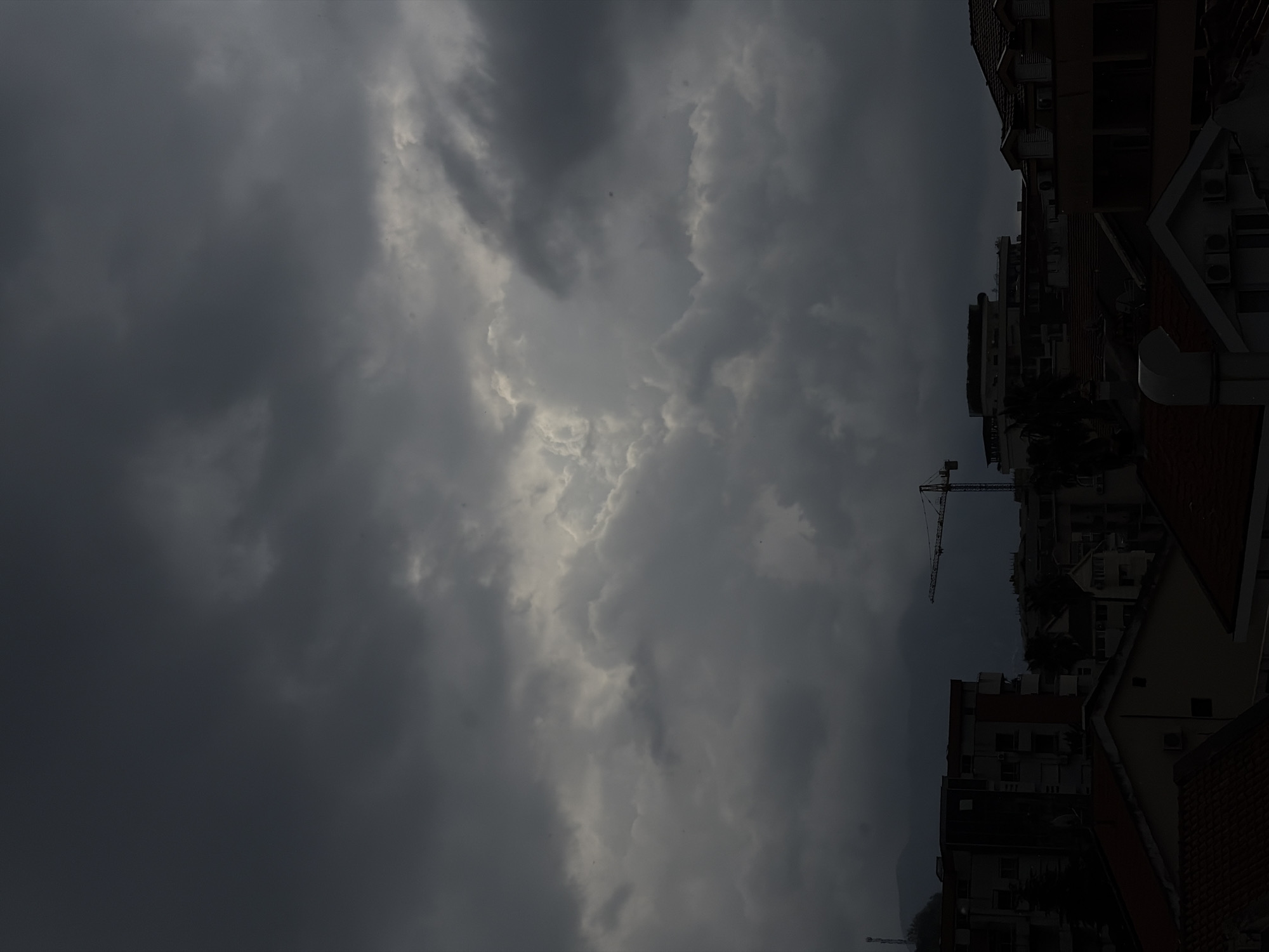 The image, when viewed in a landscape orientation, depicts a dramatic and heavily overcast sky above an urban setting. Dark, voluminous clouds dominate the upper two-thirds of the frame, showcasing various shades of grey, from deep charcoal to lighter, more ethereal patches where light appears to be breaking through. This central bright area within the clouds creates a stark contrast, suggesting either the sun attempting to pierce the cloud cover or reflecting off the upper layers of a thick cloud bank.
In the lower right portion of the image, the tops of several buildings are visible, characterized by their multi-story structures and varying roof styles, including red-tiled and flatter profiles. Air conditioning units are discernible on some building facades. A prominent construction crane extends upwards from behind these buildings, silhouetted against the dark sky, indicating ongoing development in the area. In the far background, faint outlines of what appear to be mountains or hills are vaguely visible through the hazy atmosphere. The overall scene conveys a sense of an impending storm or a day with extremely heavy cloud cover, suggesting late afternoon or early evening. The dark, brooding clouds evoke a somber or dramatic mood. No visible text is present in the image. The architectural style and presence of a construction crane are consistent with a developing urban environment like Budva, Montenegro.
