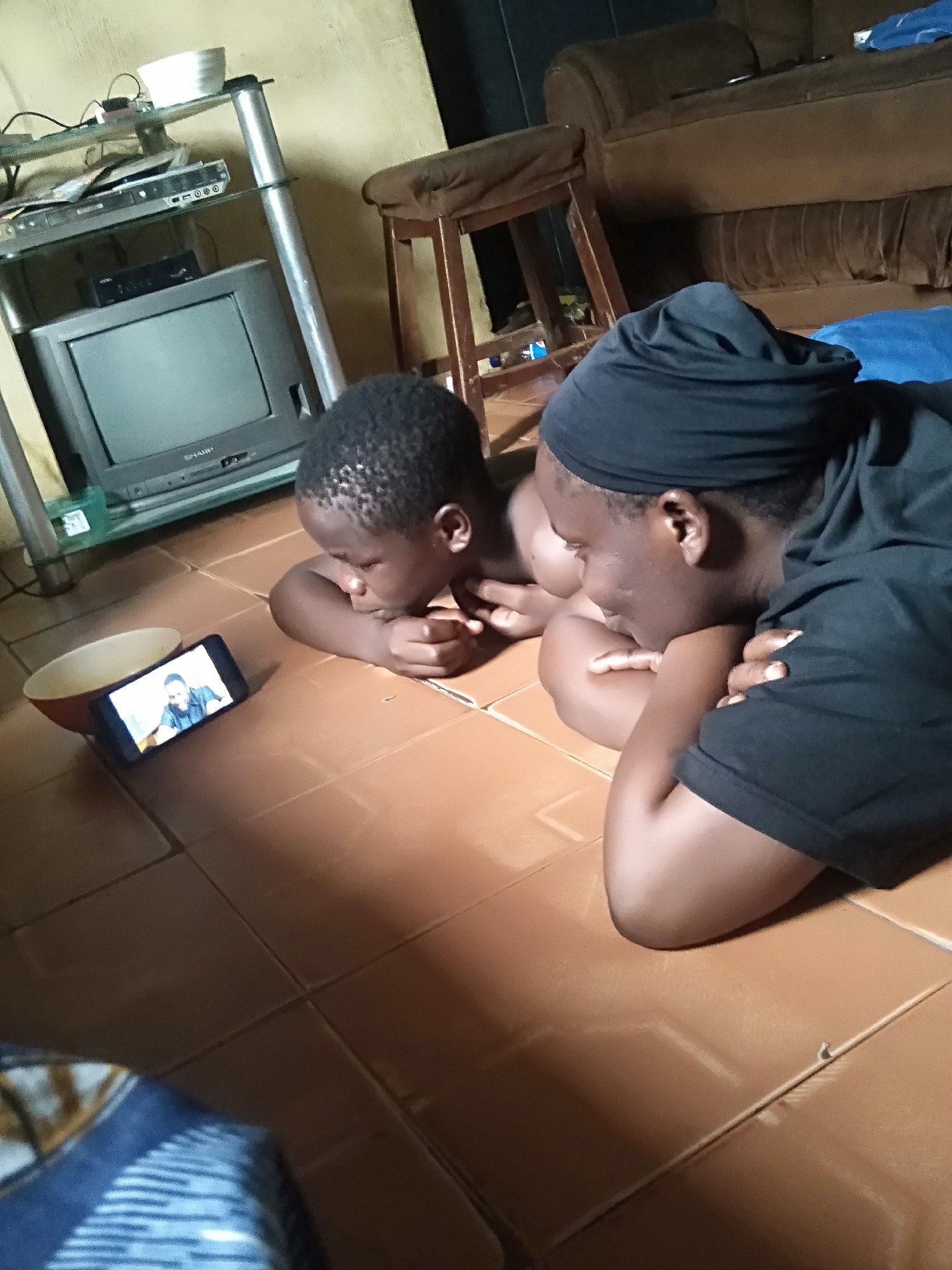 A medium shot shows two people laying on a tiled floor, watching a mobile phone. One person is a child with short, dark hair. The other is an adult with a dark head wrap and a dark shirt. Their faces are directed towards the phone, which is placed near a bowl on the floor. The screen of the phone displays a man’s face. The background includes furniture such as a TV on a metal stand, a wooden stool, and a brown couch. The scene is indoors. I cannot confirm the location in the city of Shangisha, Nigeria from this image.