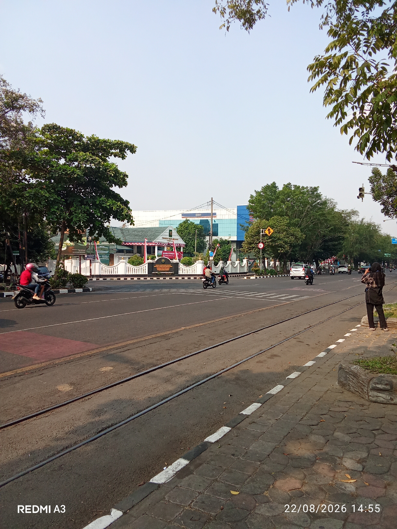 This is an eye-level, outdoor, daytime, vertical shot of a street scene in Surakarta, Indonesia, on August 22, 2026, at 2:55 PM. The foreground features a sidewalk with interlocking stone pavers and a curb with alternating white and black painted sections, bordering a street with tram tracks. The street is asphalt, with a red painted section to the left.
In the middle ground, several vehicles are visible. A motorcycle carrying two individuals, one wearing a red jacket and helmet, travels on the left side of the road. Further down the street, other motorcycles and cars are in motion. To the left, a building with a white fence and red and white banners appears to be a public institution, possibly a court or government office, with a large green tree in front. In the background, a modern building with blue glass windows is visible, along with more trees and a traffic light.
On the right side of the sidewalk in the foreground, a woman stands facing away from the camera, holding a phone up to her face as if taking a picture or video. She is wearing a dark headscarf and a dark jacket over dark pants. The sky is clear and light blue, with the tops of trees and branches visible in the upper portion of the frame.