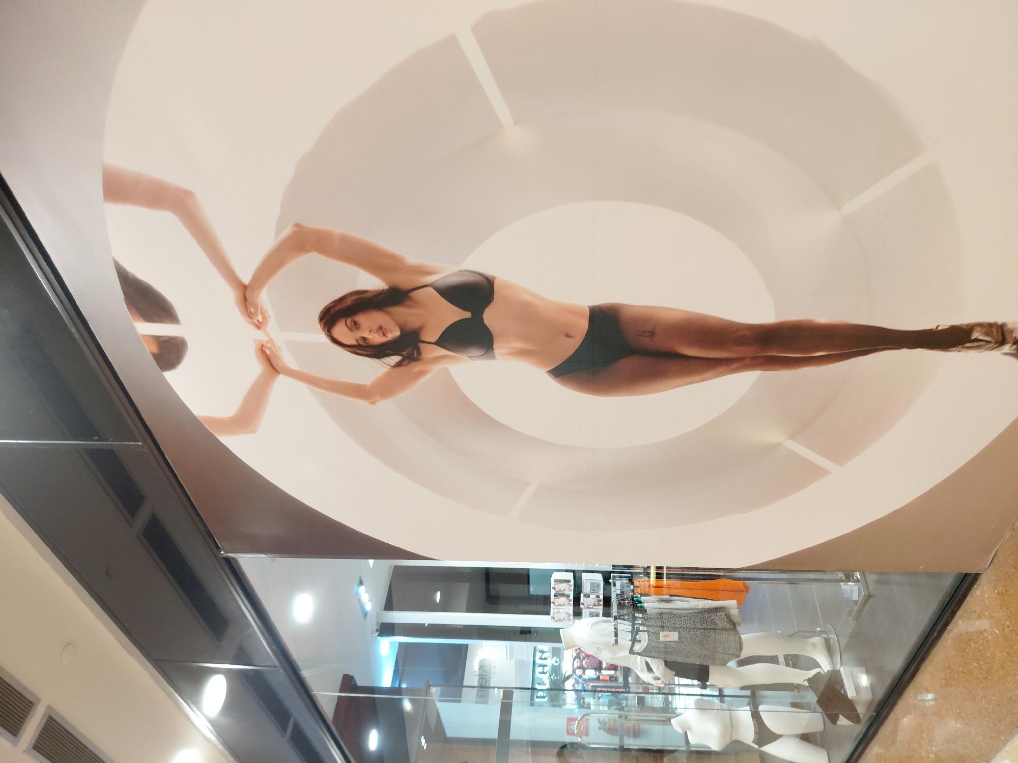 A large, full-body advertisement of a woman wearing a black bra and panties is displayed on a white wall in what appears to be a shopping mall. The woman is posing with her arms raised. To the left of the advertisement, there is a store window with mannequins displaying clothing. The store appears to be a clothing retailer. The overall scene is brightly lit, suggesting an indoor environment.