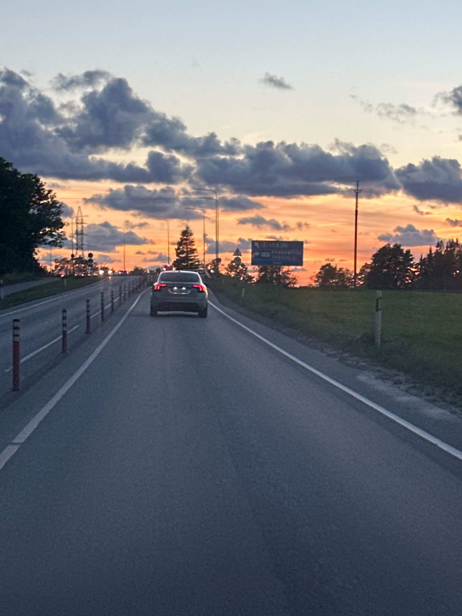 A dark-colored sedan is traveling on a paved road, with the rear of the vehicle in the foreground. The road is lined with white markings and intermittent reflective bollards on the right side. The sky above is a gradient of pale blue to vibrant orange and pink, illuminated by a setting sun. Dark, cumulonimbus clouds are scattered across the sky, some reflecting the sunset's colors. On the right side of the road, a grassy verge is visible, with scattered trees and a signpost indicating directions. The signpost has blue text and symbols. On the left side of the road, there are trees, power lines, and other vehicles in the distance. The scene is captured at dusk on a road, possibly on the outskirts of Hüüru, Estonia, given the signage.