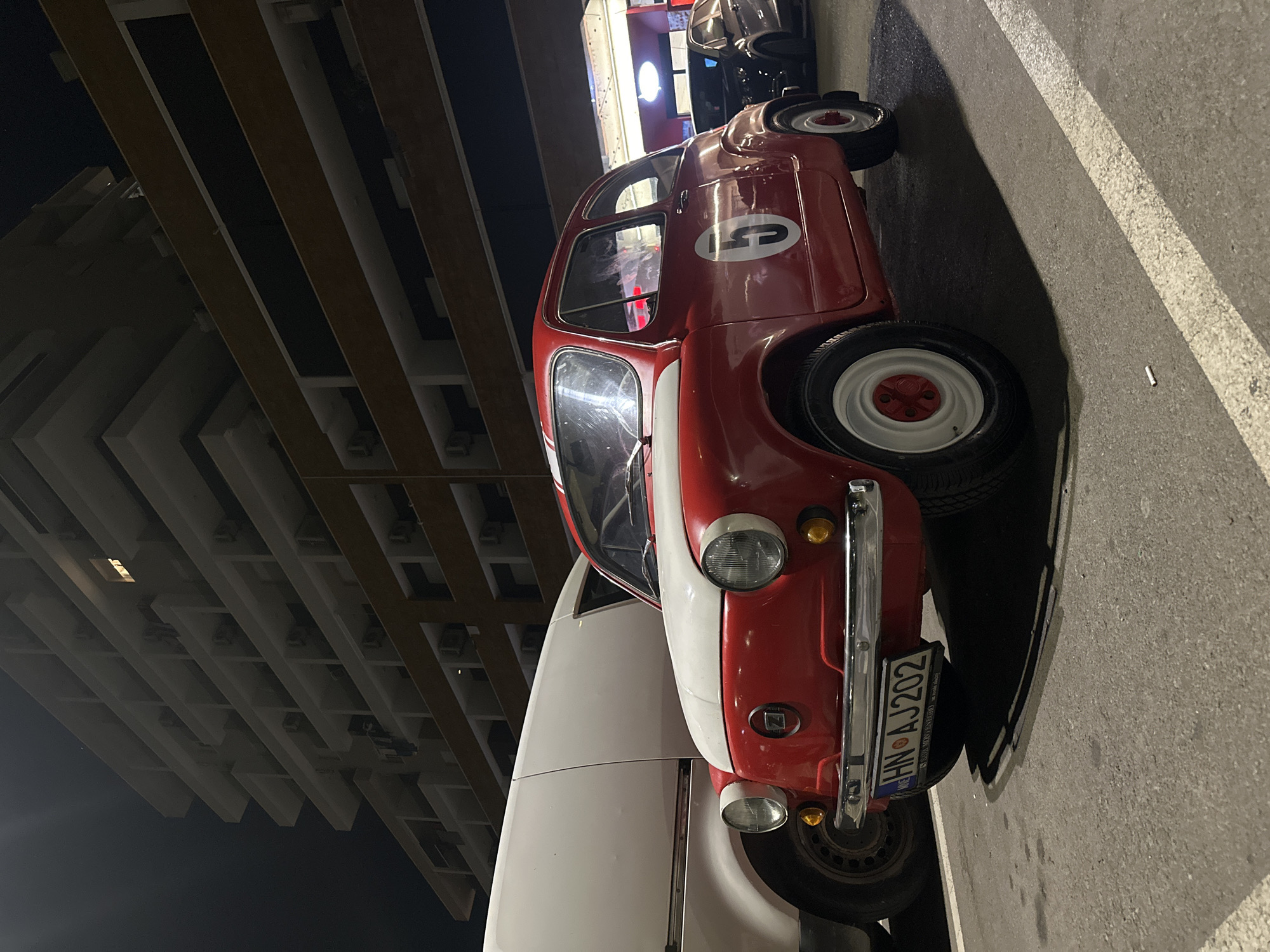 A red and white vintage Fiat 600 is parked at night in a parking lot in Budva, Montenegro, with a large apartment building in the background. The car is facing the viewer and is adorned with white racing stripes on the hood and a large white number "5" on its side. A white van is parked to the left of the Fiat. The apartment building features multiple balconies with air conditioning units and some lit windows. The street is paved, and a white line marks a parking space. The sky is dark, indicating nighttime.