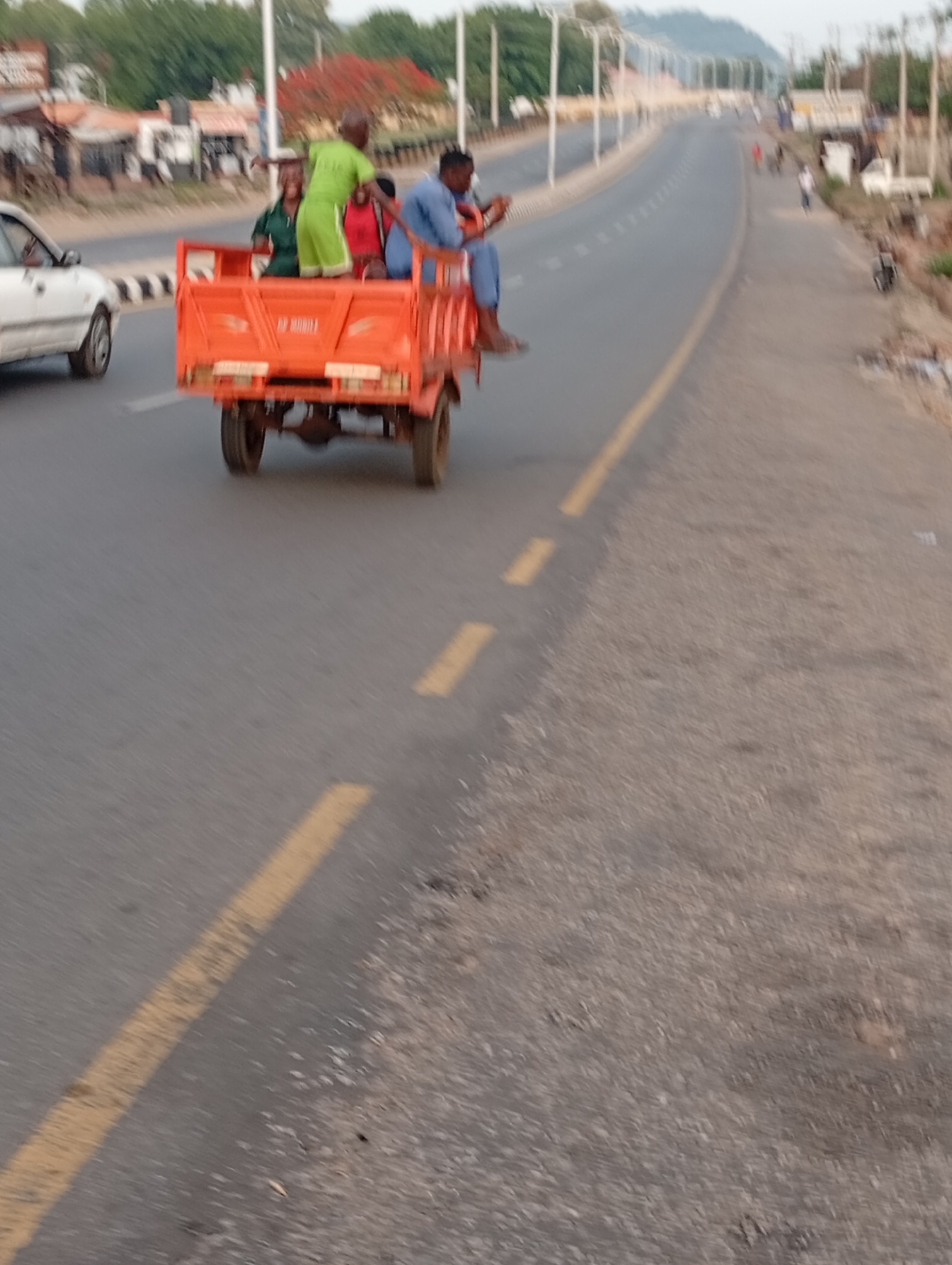 An orange open-carriage motor vehicle with four passengers moves along a divided highway in Adankolo, Nigeria. The highway has a yellow dividing line. The passengers include a man wearing a green outfit, a man wearing a blue outfit, and two other people. A white car is in the adjacent lane. Buildings and trees line the road, and overhead lighting is visible. Other pedestrians can be seen in the distance. The road surface is visible, showing some wear and debris.
