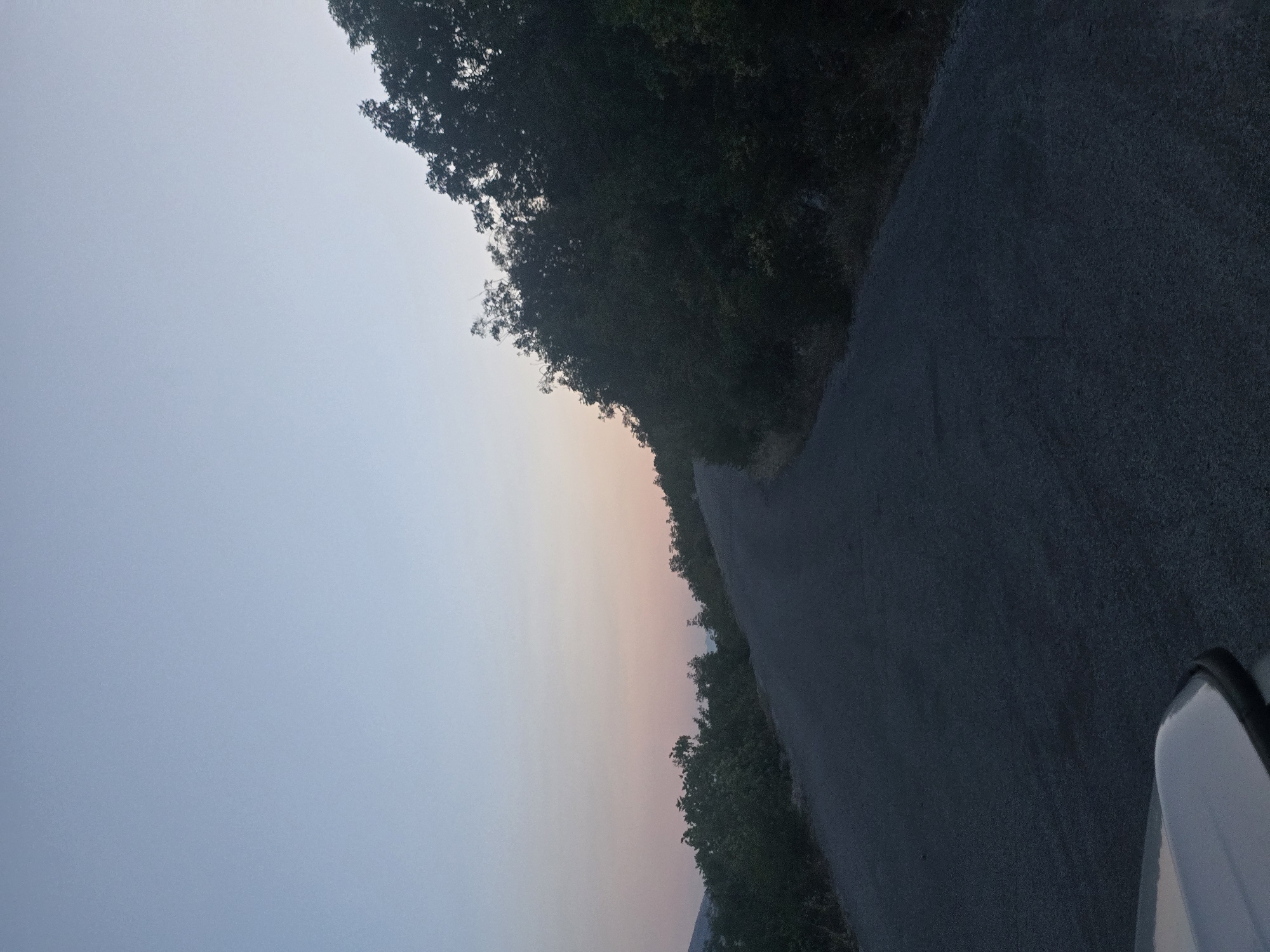 The image depicts a winding asphalt road ascending a hillside under a twilight sky. Dense, dark green foliage lines the right side of the road, forming a thick forest. On the left side, the foliage is sparser and appears further in the distance, with a hint of mountains visible on the horizon. The sky transitions from a pale blue at the top to soft hues of pink and orange near the horizon, indicating either sunrise or sunset. The foreground shows the hood of a light-colored vehicle on the left side of the frame, suggesting the image was taken from a car. The road surface is textured and appears to be in good condition. The scene is devoid of any human presence or activity. The location is specified as Klopot, Montenegro.