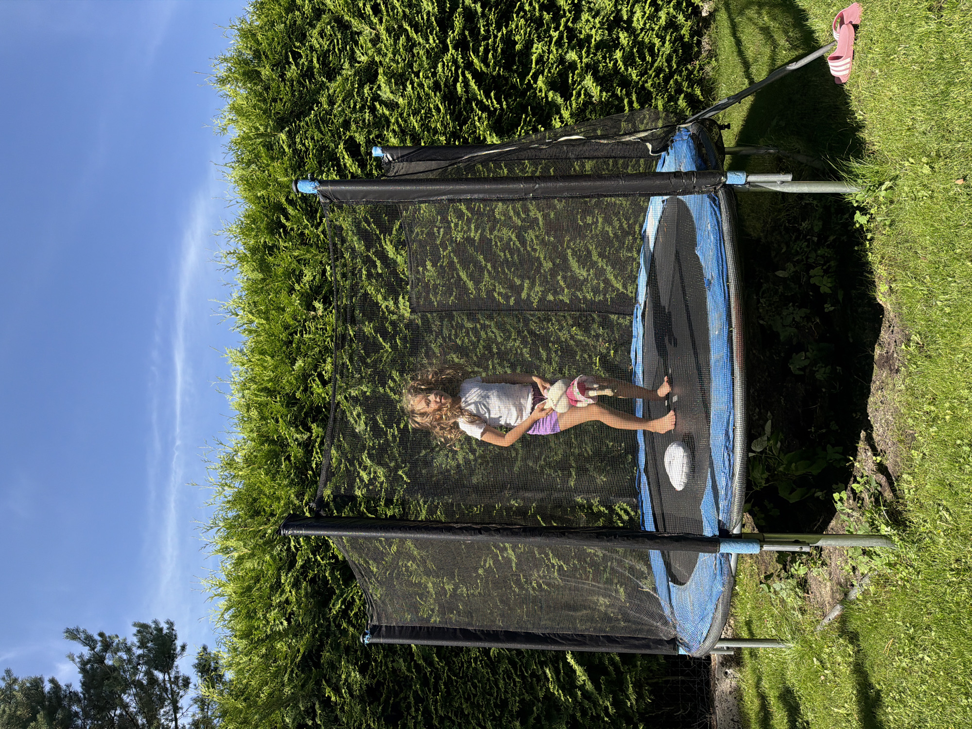A young girl with curly blonde hair is standing on a trampoline in a backyard. She is wearing a white t-shirt and purple shorts, and holding a stuffed animal. The trampoline has a black safety net and a blue rim. Behind the trampoline is a dense green hedge. The sky is blue with some wispy clouds. The ground is covered in green grass. A pair of pink flip-flops are on the grass to the right of the trampoline. This scene is likely in the city of Baloži, Latvia.