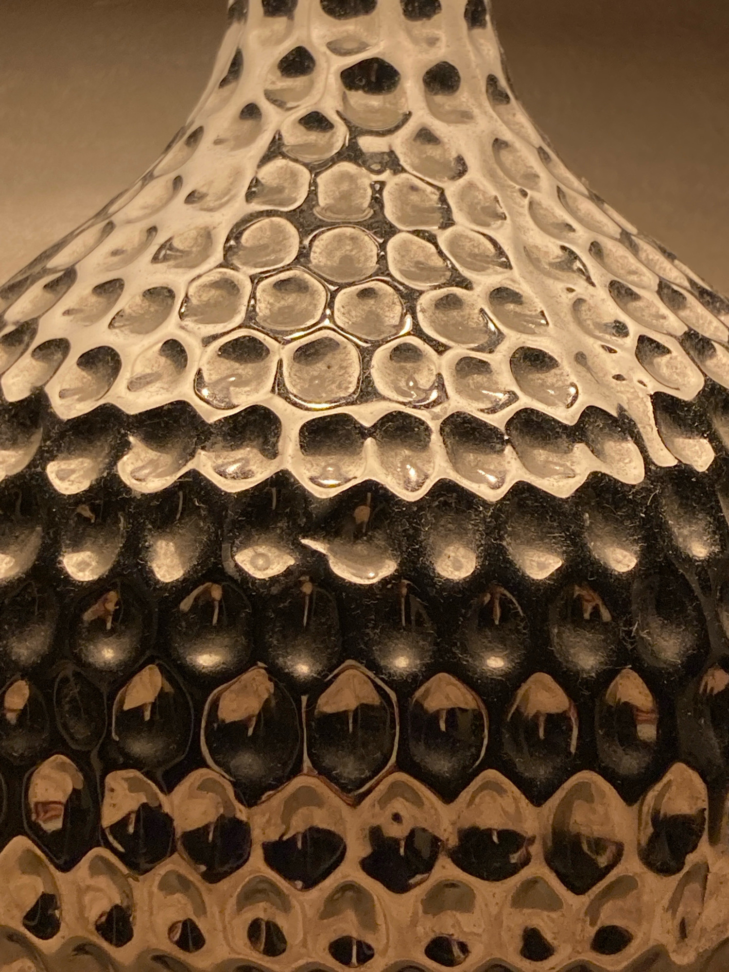 The image is a close-up, angled shot of a textured object, likely a vase or decorative piece. The surface features a repeating pattern of hexagonal indentations, resembling a honeycomb or reptile scales. The object transitions in color from a lighter, possibly white or cream at the top, to a darker, black or deep grey in the middle section, and then to a warm, metallic brown or copper at the bottom. The indentations create a play of light and shadow, with highlights reflecting off the smooth inner surfaces of the cavities and darker tones in the recessed areas. The texture is uneven and organic, suggesting it is handmade or has an aged appearance. The background is a blurred, neutral, warm-toned surface, indicating an indoor setting with soft lighting. There is no indication of the city of Samara, Russia, in the image.