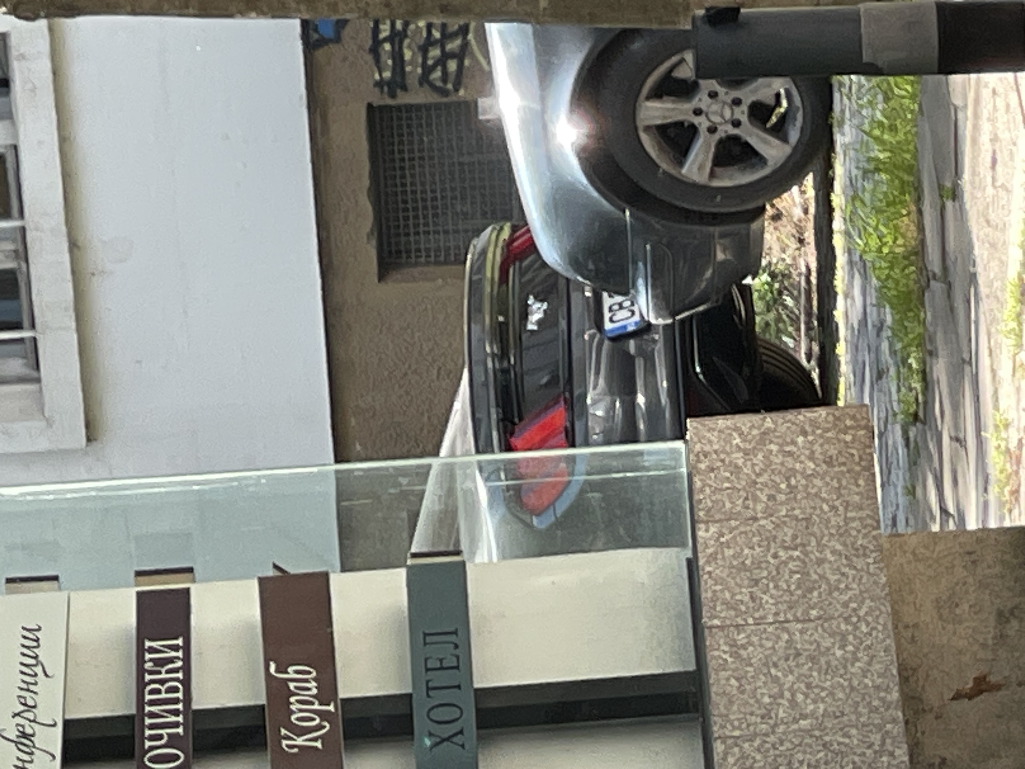 A rear view of a parked gray Mercedes-Benz is framed by a window display of a business that reads "ХОТЕЛ" in Bulgarian Cyrillic, indicating "HOTEL" in English. Further descriptions of the business mention "Кораб", "ОЧИВКИ", and "Конференции" which may relate to hotel amenities or services. The location is possibly in Sofia, Bulgaria as the license plate on the car contains "CB" which is a regional code for Sofia. The car is parked on the side of a street next to a green area that includes grass and trees. The building behind the car is white with a dark gray window grate. A light gray concrete pillar is on the right-hand side of the shot.