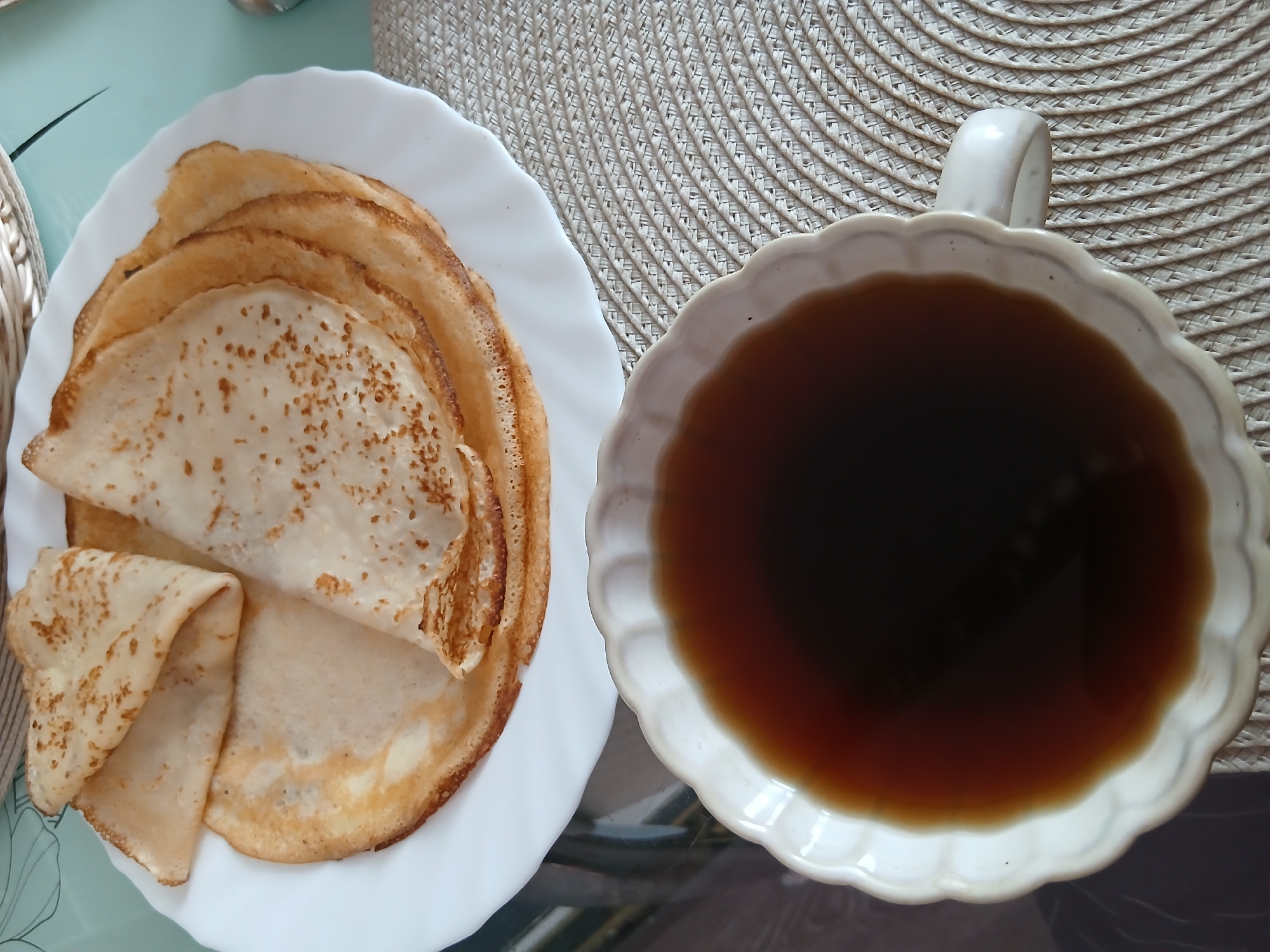A close-up, overhead shot captures a white plate of crepes and a white mug of dark liquid. The crepes are a golden-brown, with some slightly darker spots, and are stacked on the plate. One crepe is folded into a triangle, resting on top of the stack. The mug, with a scalloped edge, is positioned in the foreground, filled with a dark, amber-colored liquid that appears to be tea or coffee. The mug sits on a woven placemat with a light beige color and a diagonal pattern. The background is blurred, but suggests a table setting, possibly in a home environment in Chaikovsky, Russia.