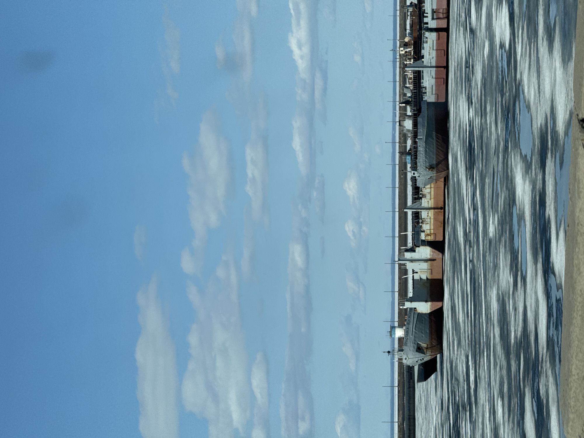 The image shows a view of the Kama River in Chaikovsky, Russia. Several decommissioned river vessels, including "PRISTAVKA-VOLZHSKIY", are docked along the frozen riverbank. The sky is blue with scattered clouds. The frozen river surface displays a textured pattern of ice and water. The riverbank is sandy. Power poles stand along the bank.