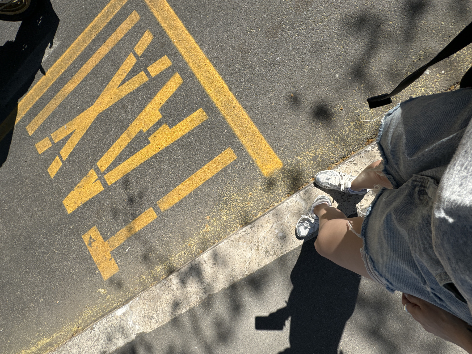 The image shows a partial view of a person standing next to a "TAXI" marking painted in yellow on asphalt. The person is wearing denim shorts and white sneakers. Part of a car is visible in the upper left corner, casting a shadow. The environment appears to be a street in an urban setting. The specific location in Rome, Italy, is not identifiable from the given image.